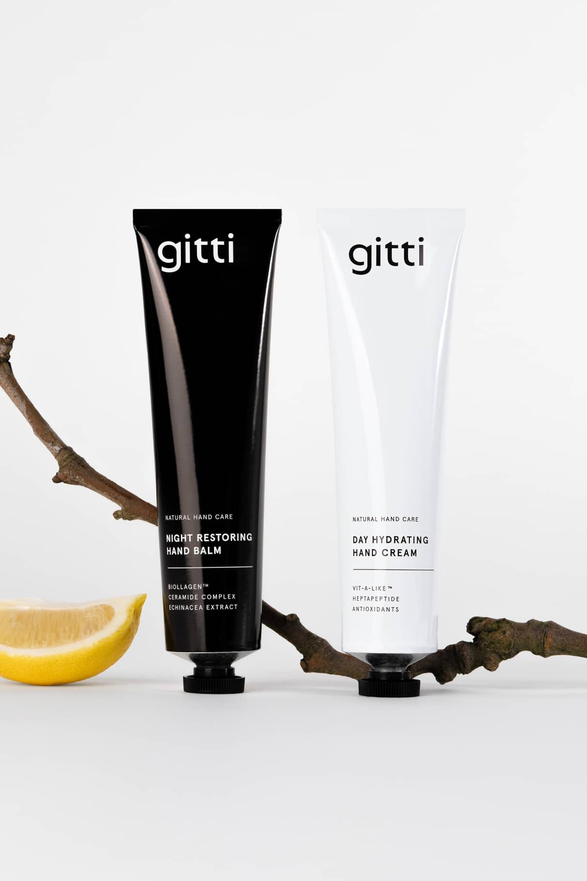 Zwei gitti 24 Hour Hand Therapy Tuben - eine schwarze (Night Restoring Hand Balm) und eine weiße (Day Hydrating Hand Cream) - stehen aufrecht neben einem Baumzweig und einer Zitrone und bieten die 24-Stunden-Pflege für weiche Hände.
