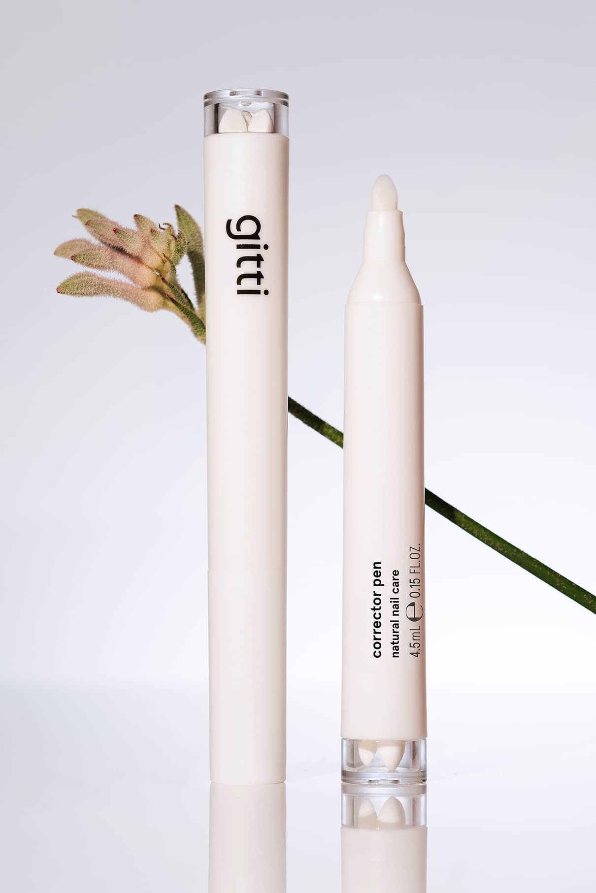 Zwei weiße Korrekturstiftbehälter mit der Aufschrift gitti und Nagellackentferner-Stift stehen aufrecht neben einer hellrosa Blume auf einer spiegelnden Fläche und heben Werkzeuge für ein makelloses Finish vor einem neutralen Hintergrund hervor.
