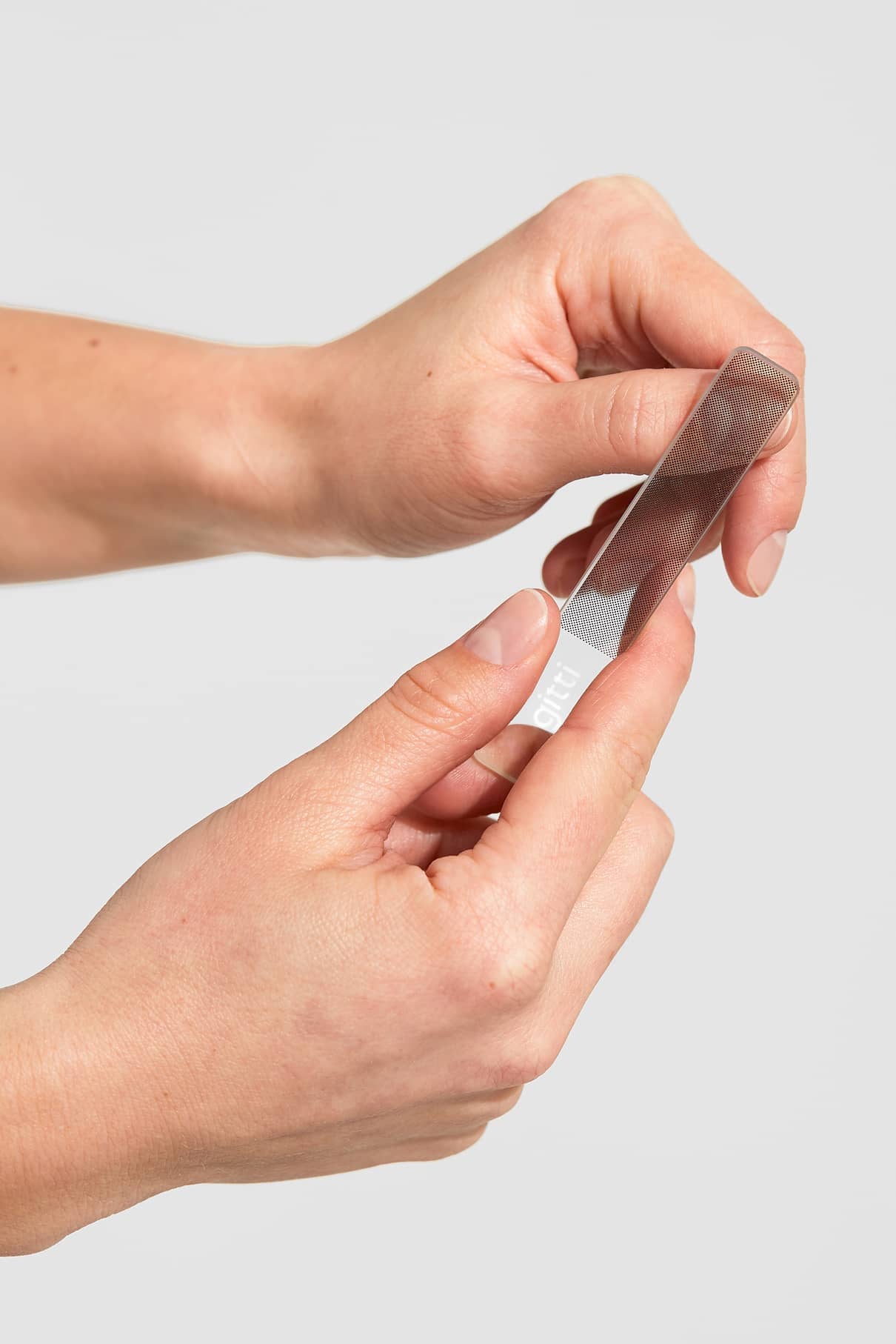 Eine Person hält eine Metallpinzette in der rechten Hand und justiert mit der linken Hand die Spitze - möglicherweise nach der Verwendung eines Nagelbuffers für glänzende Nägel - vor einem glatten Hintergrund.