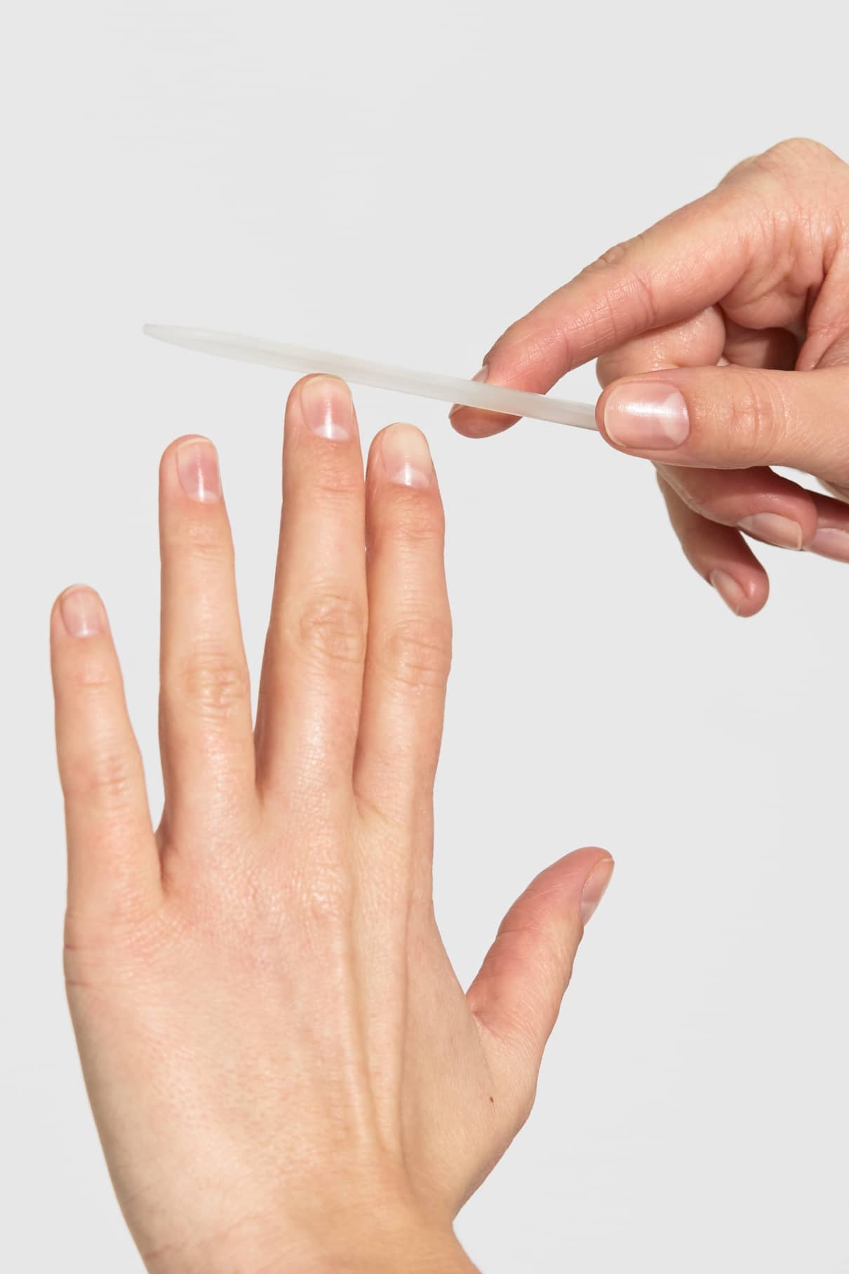 Eine Person benutzt eine Nagelfeile, um die Fingernägel ihrer linken Hand vor einem schlichten hellgrauen Hintergrund zu formen.