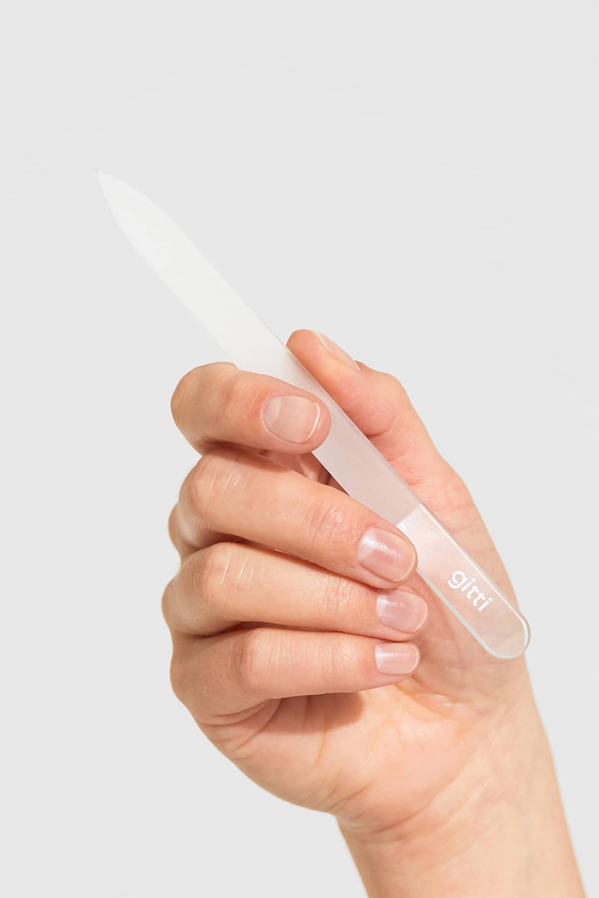 Eine Hand, die eine gläserne Nagelfeile aus Glas mit dem Markennamen auf dem Griff hält, vor einem schlichten hellen Hintergrund.