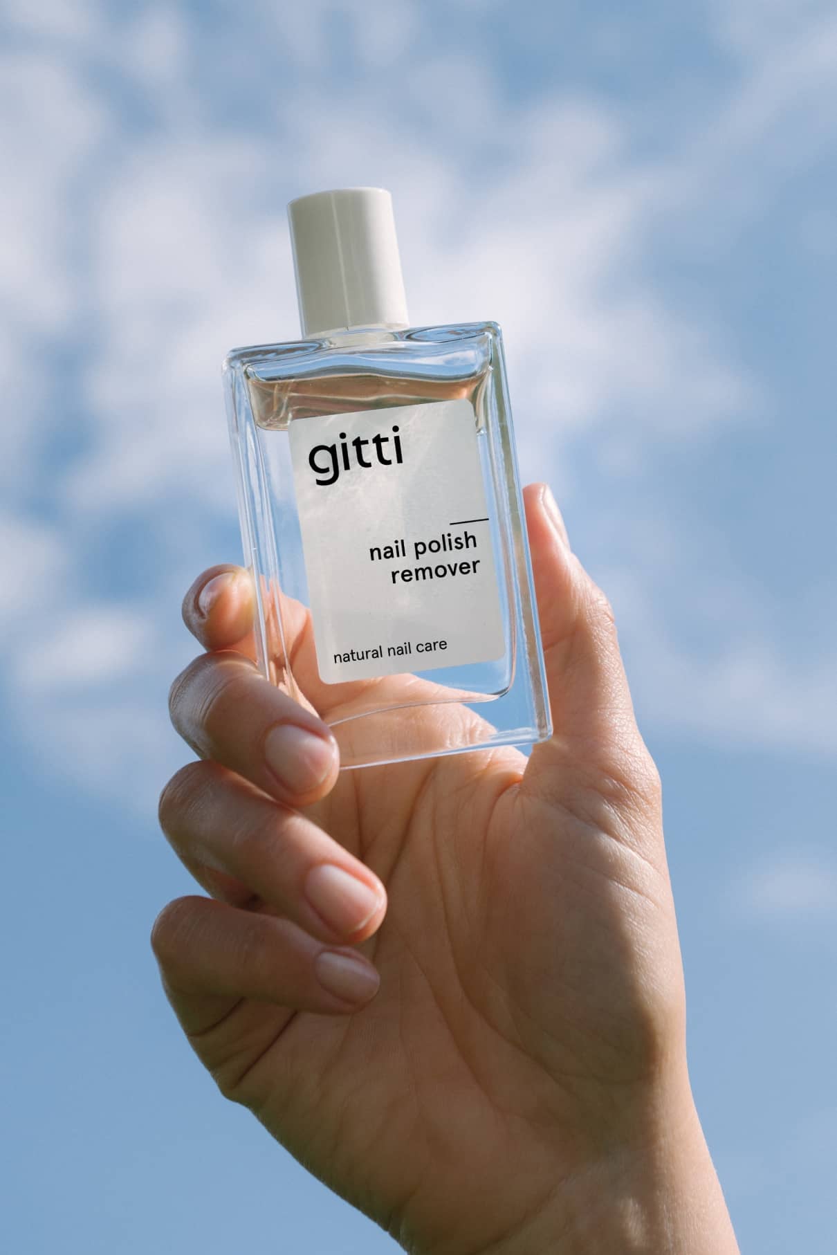 Eine Hand hält eine Klarglasflasche mit der Aufschrift Nagellackentferner für Naturnagelpflege mit angenehmem Duft vor einem Hintergrund mit blauem Himmel.