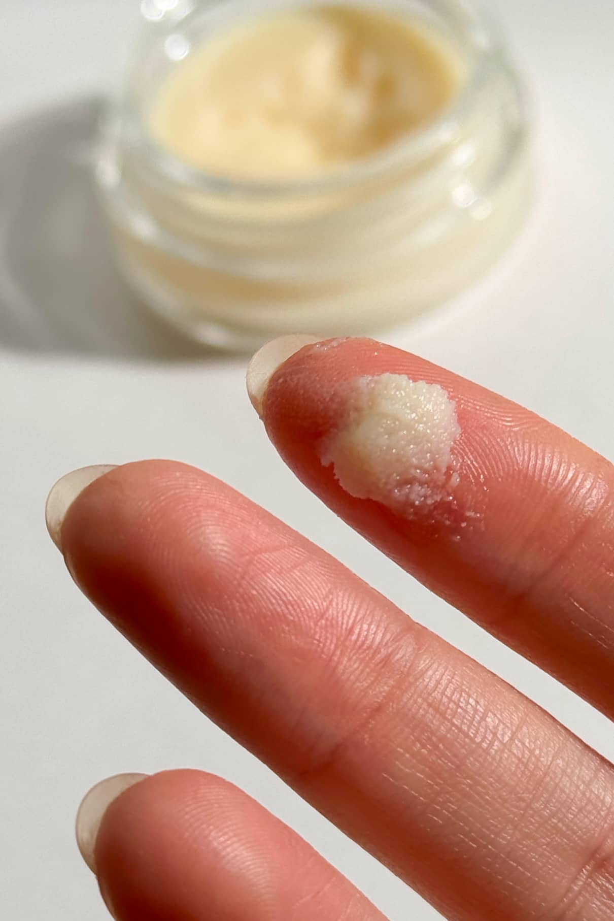 Nahaufnahme von Fingern, die eine kleine Menge Nagellackentferner Balsam halten, mit einem geöffneten Behälter des cremig-beigen Produkts im Hintergrund.
