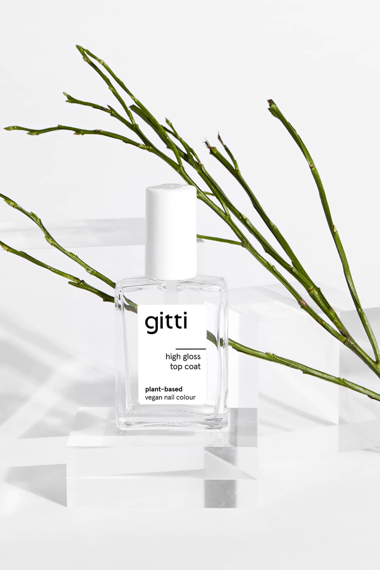 Eine durchsichtige Flasche mit High Gloss Top Coat Nagellack steht auf transparenten Blöcken, eingerahmt von grünen Zweigen im Hintergrund.