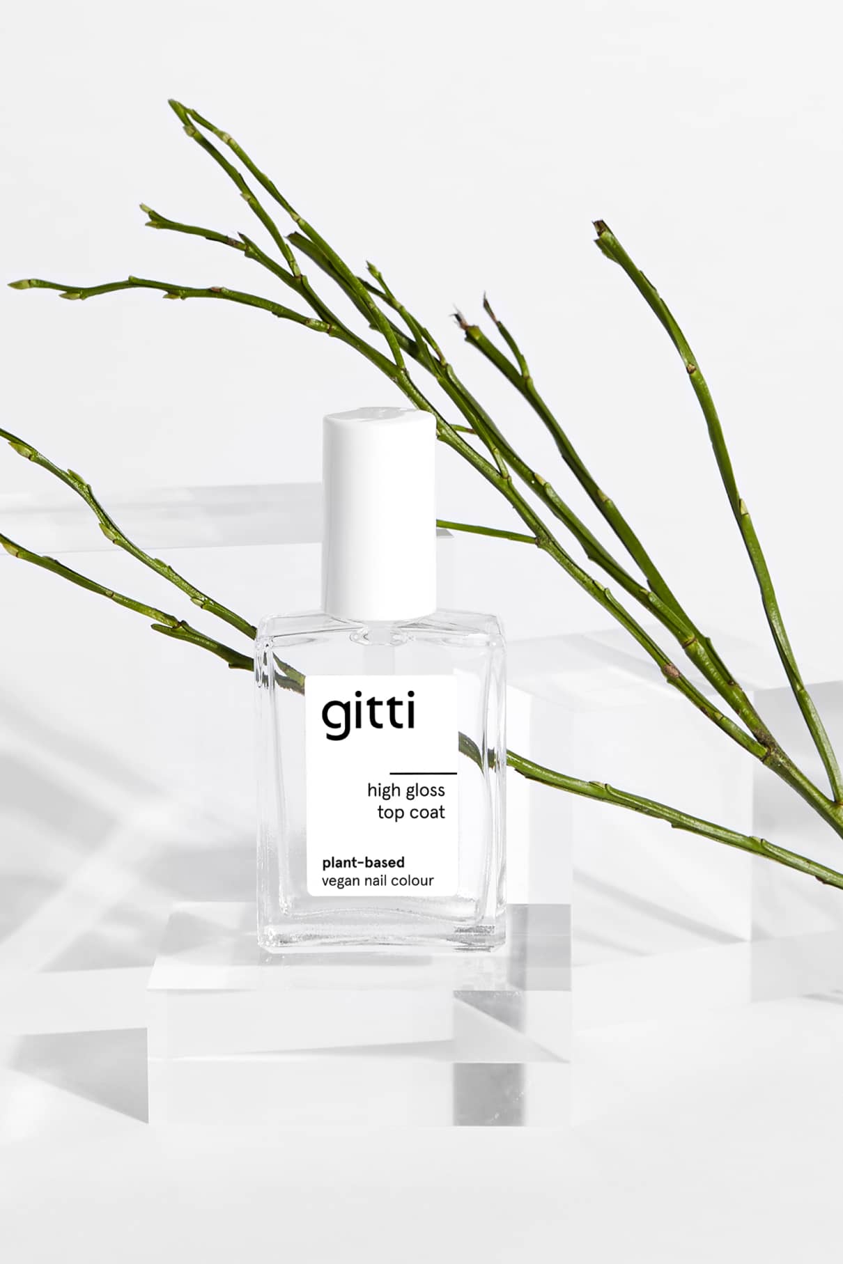 Eine durchsichtige Flasche mit High Gloss Top Coat Nagellack steht auf transparenten Blöcken, eingerahmt von grünen Zweigen im Hintergrund.