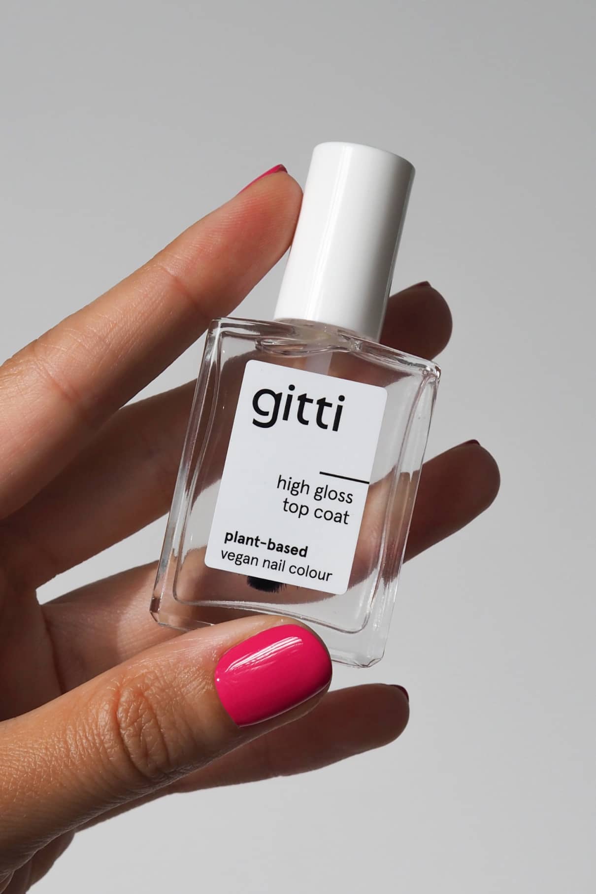Eine Hand mit rosa lackierten Nägeln hält eine Flasche High Gloss Top Coat, eine pflanzliche und vegane Nagelfarbe, die ein brillantes Hochglanzfinish verspricht.