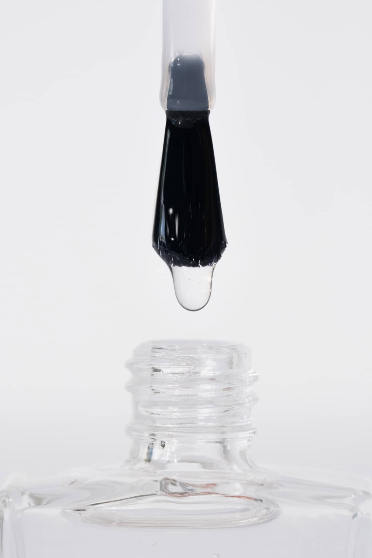 Eine Nahaufnahme zeigt den Hochglanz-Topcoat-Pinsel mit einem klaren Tropfen, der über die offene Mündung der transparenten Flasche gehalten wird, vor einem weißen Hintergrund, der das brillante Hochglanz-Finish zur Geltung bringt.