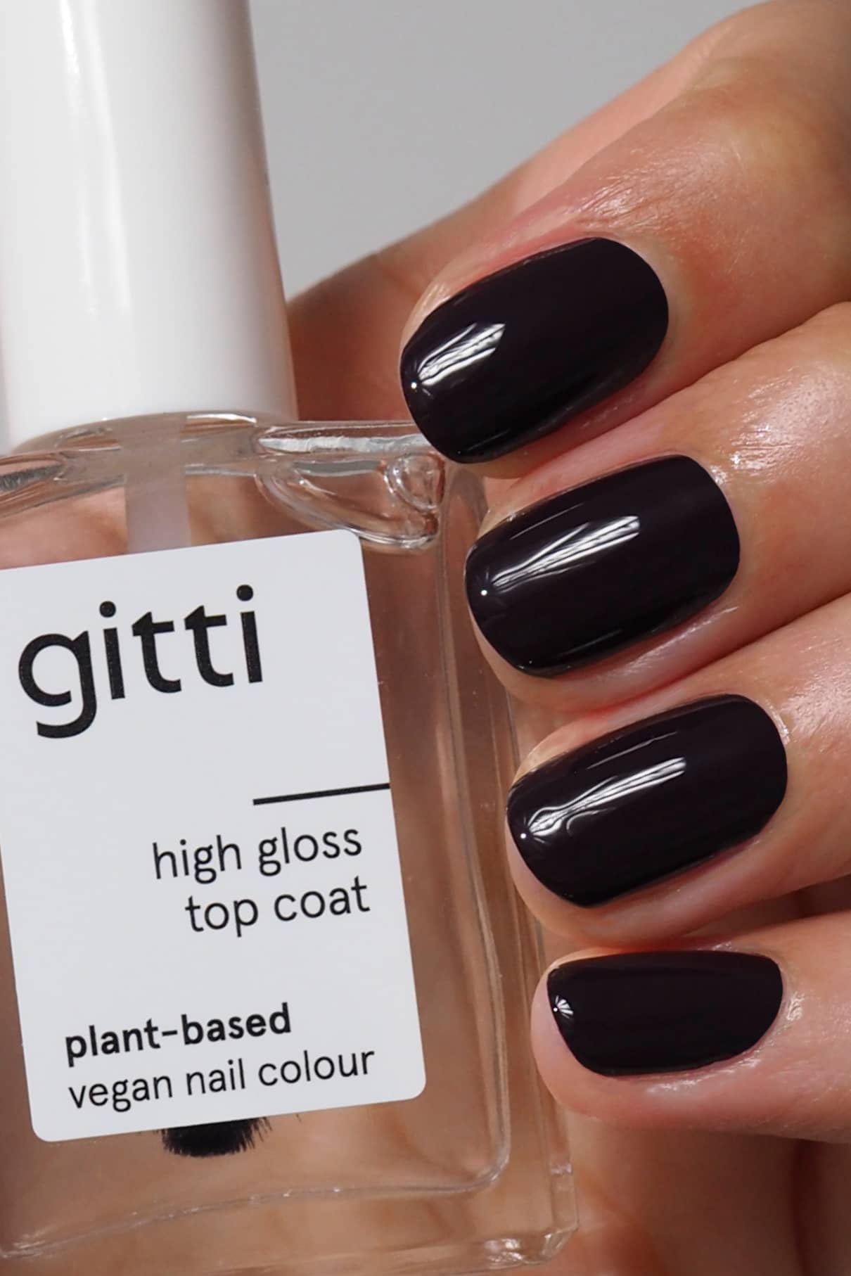 Eine Hand mit dunklen, glänzenden Nägeln hält eine Flasche mit der Aufschrift "High Gloss Top Coat", die das intensive, glänzende Finish und die pflanzliche, vegane Formel hervorhebt.