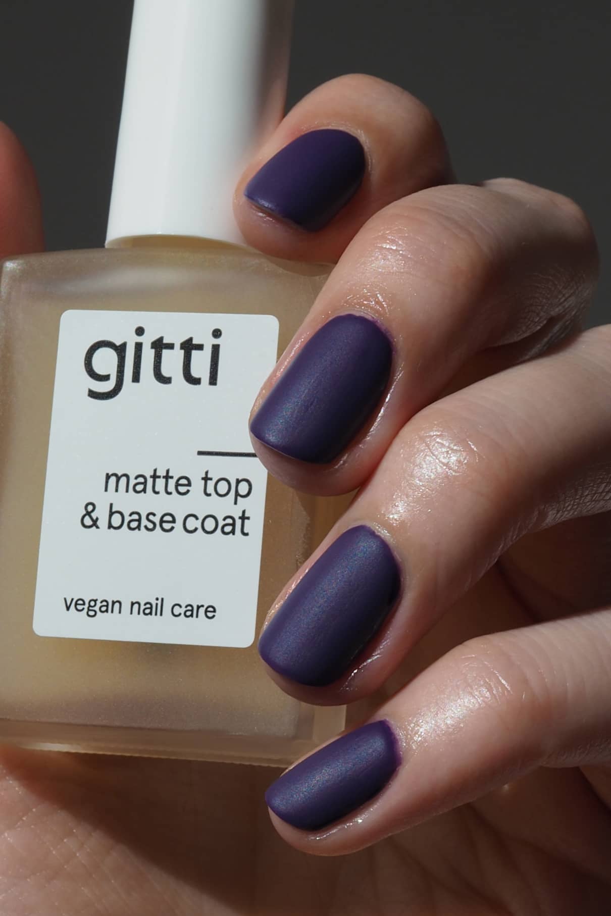 Eine Hand mit matten, dunkelvioletten Nägeln hält eine Flasche Matte Top & Base Coat - vegane Nagelpflege, die zusätzlichen Halt und ein mattes Finish in einem 2-in-1-Produkt bietet.