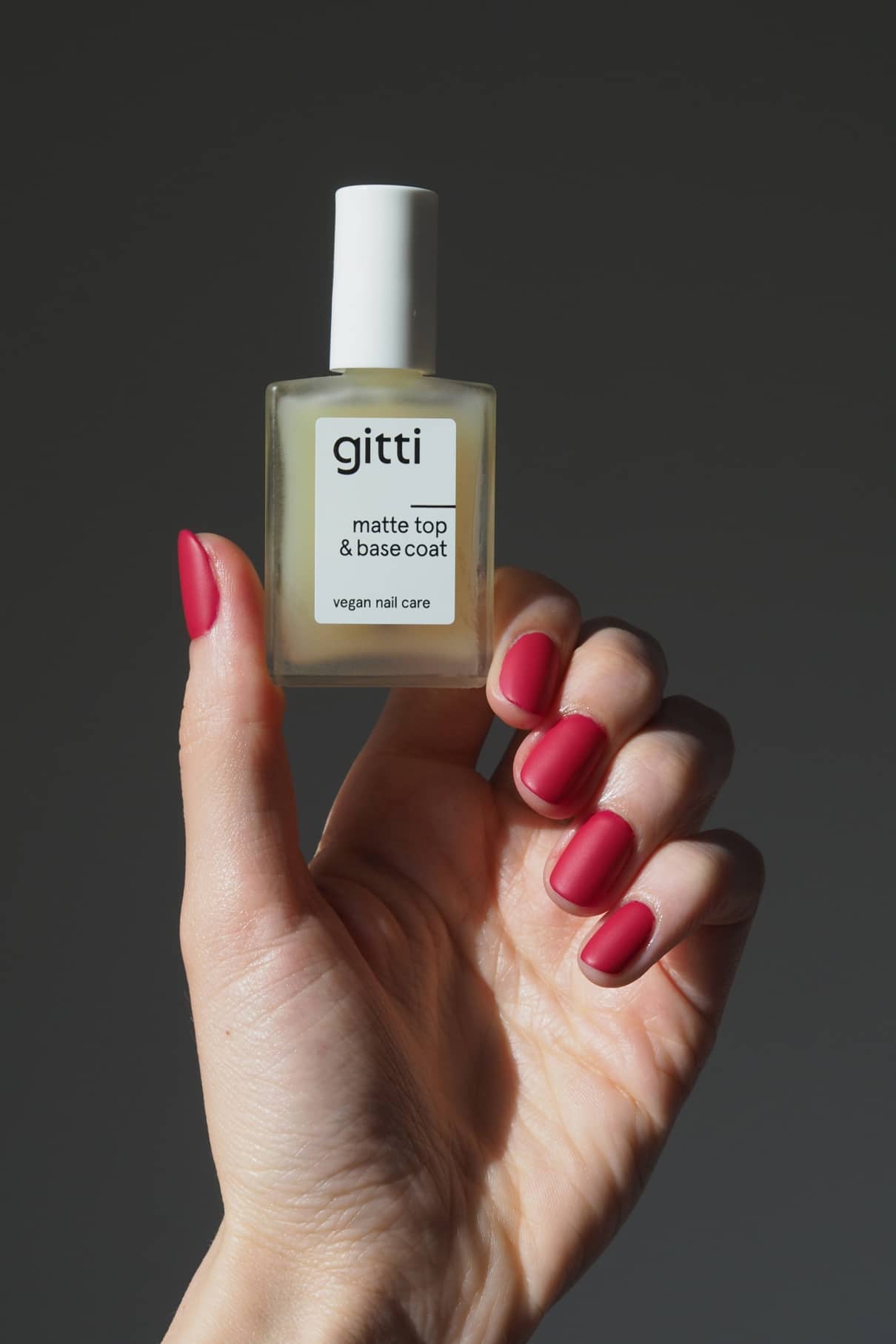 Eine Hand mit roten Nägeln hält eine Flasche Matte Top & Base Coat, ein veganes 2-in-1-Nagelpflegeprodukt für ein mattes Finish und zusätzlichen Halt, vor einem neutralen Hintergrund.
