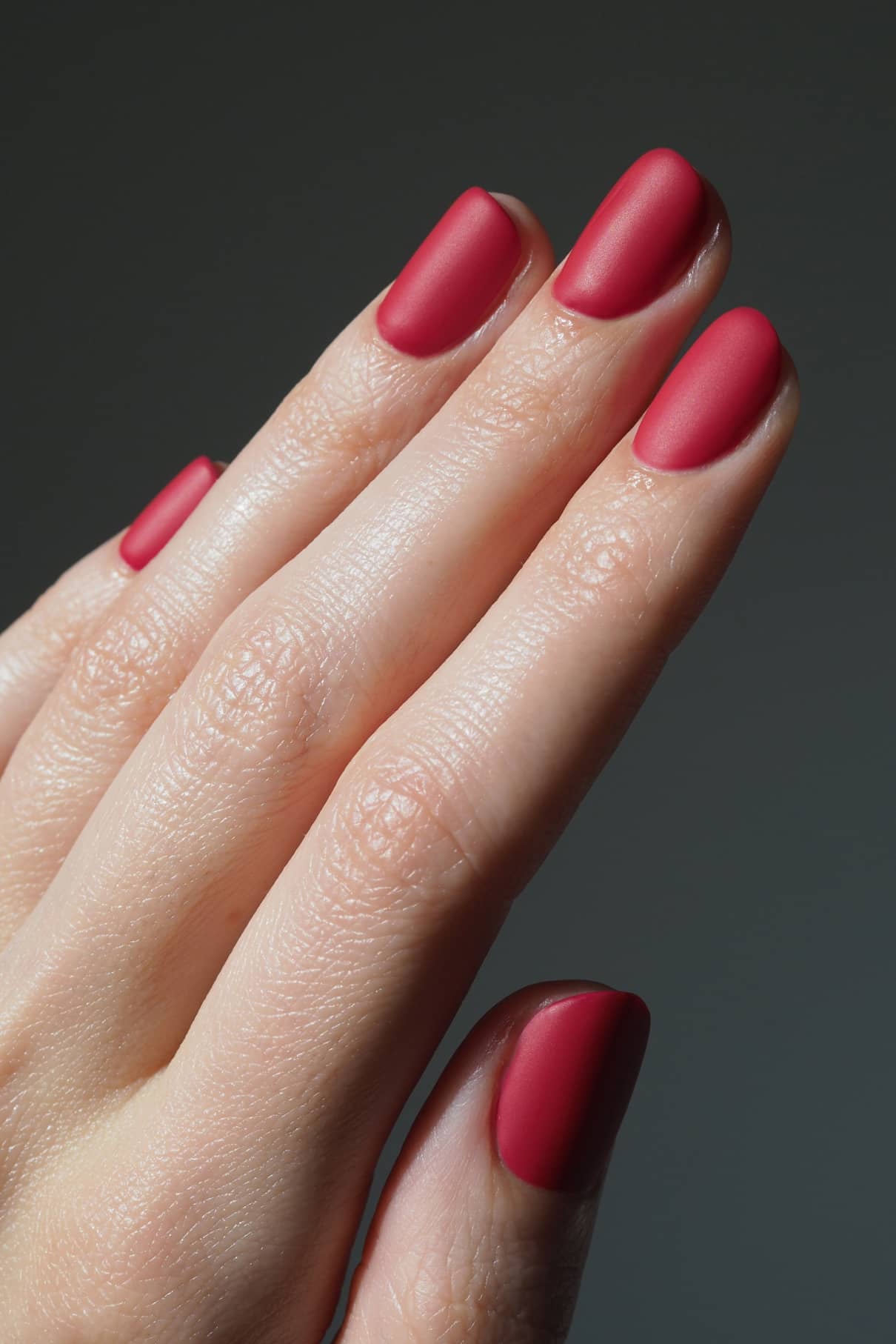 Eine Nahaufnahme einer Hand mit sauber manikürten Nägeln mit mattem rotem Lack, die das matte Finish und den zusätzlichen Halt des Matte Top & Base Coat vor einem unifarbenen Hintergrund hervorhebt.