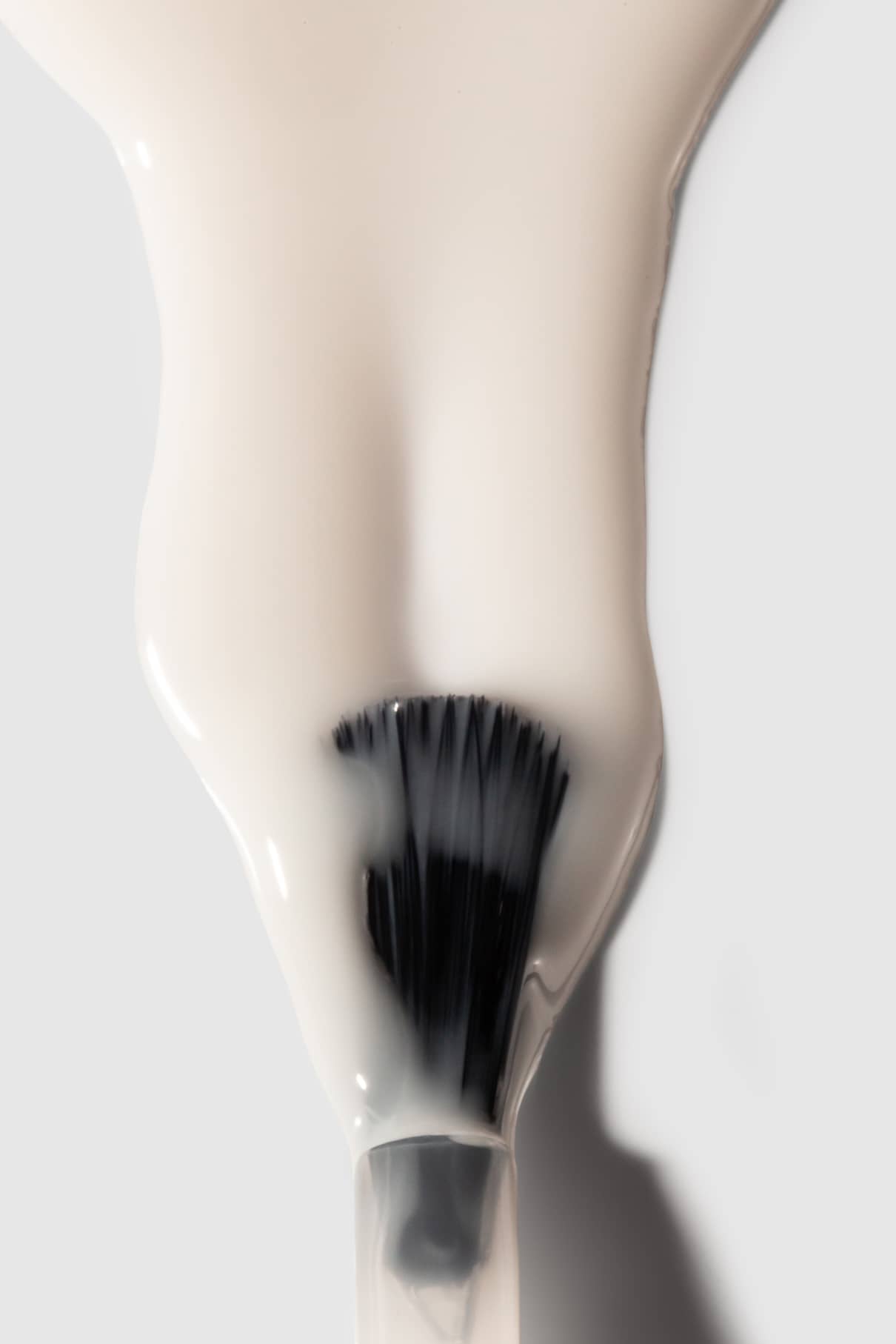 Nahaufnahme eines Pinsels, der teilweise mit Nail Glaze, einer dicken, halbtransparenten milchig-weißen Flüssigkeit, bedeckt ist, vor einem schlichten Hintergrund.