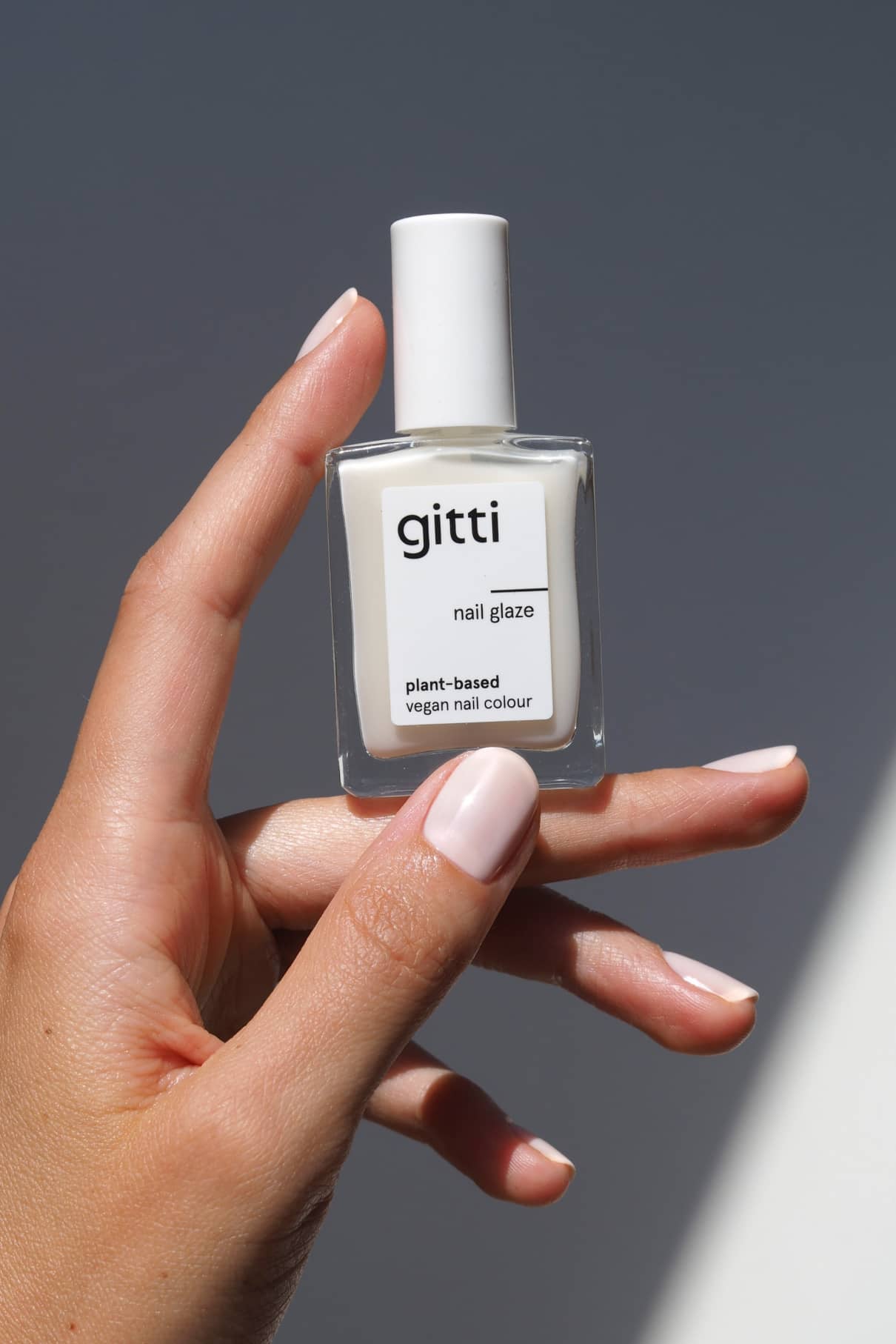 Eine Hand mit hellrosa, halbtransparenten Nägeln hält eine Nail Glaze-Flasche mit der Aufschrift pflanzlich und vegan in milchigem Weiß vor einem neutralen Hintergrund.