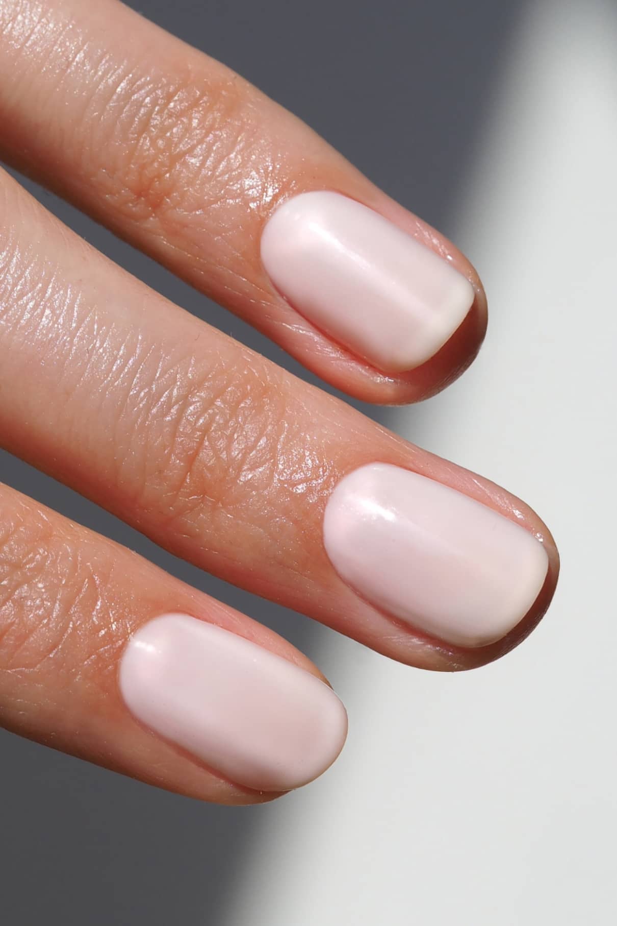 Nahaufnahme von drei Fingern mit kurzen, sauber gefeilten Nägeln, die mit Nail Glaze lackiert sind, einem glänzenden, halbtransparenten, milchigen Weiß mit einem Hauch von zartem Rosa.