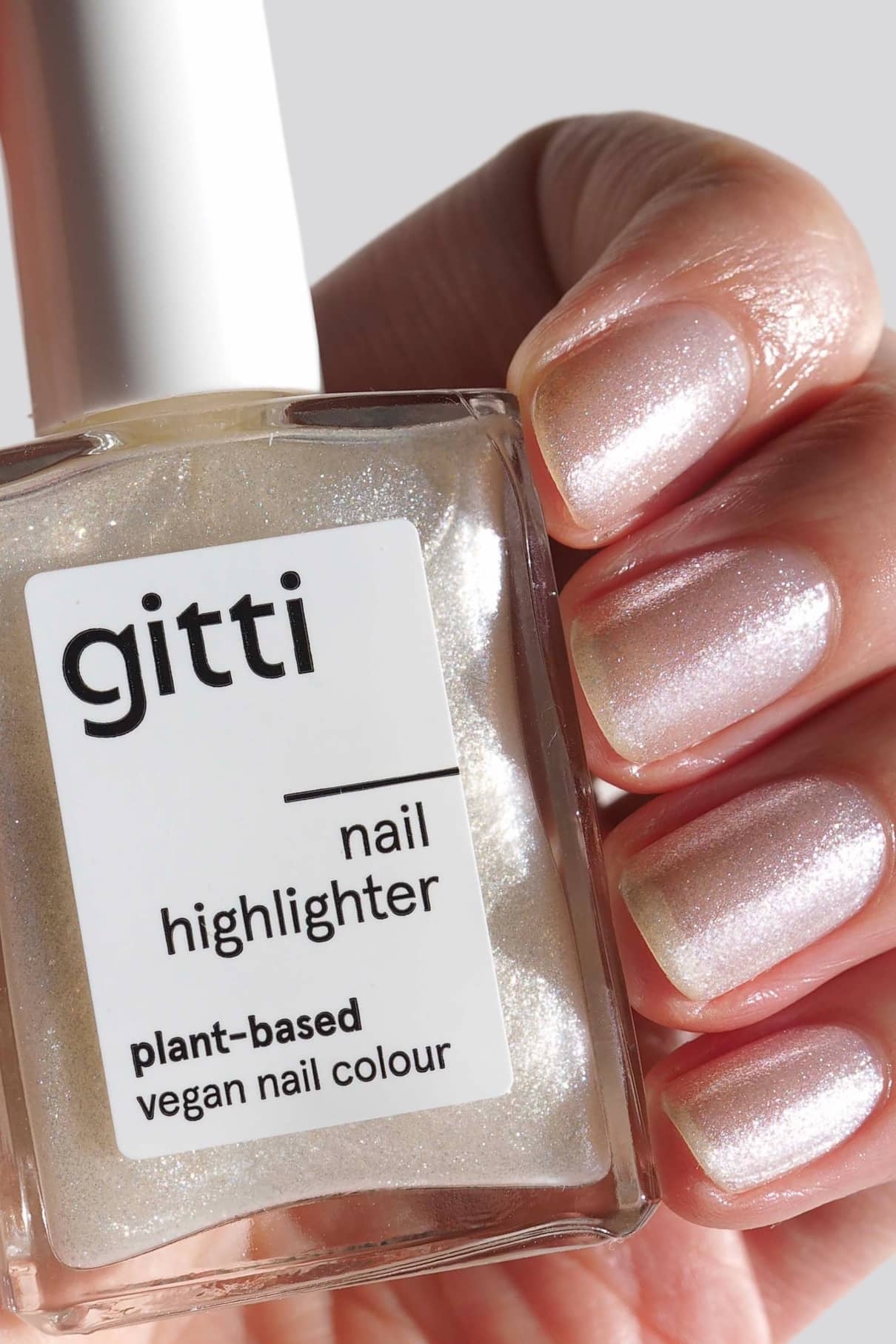 Eine Hand mit schimmernd lackierten Nägeln in zartem Ton hält eine Flasche Nail Highlighter - pflanzliche, vegane Nagelfarbe.