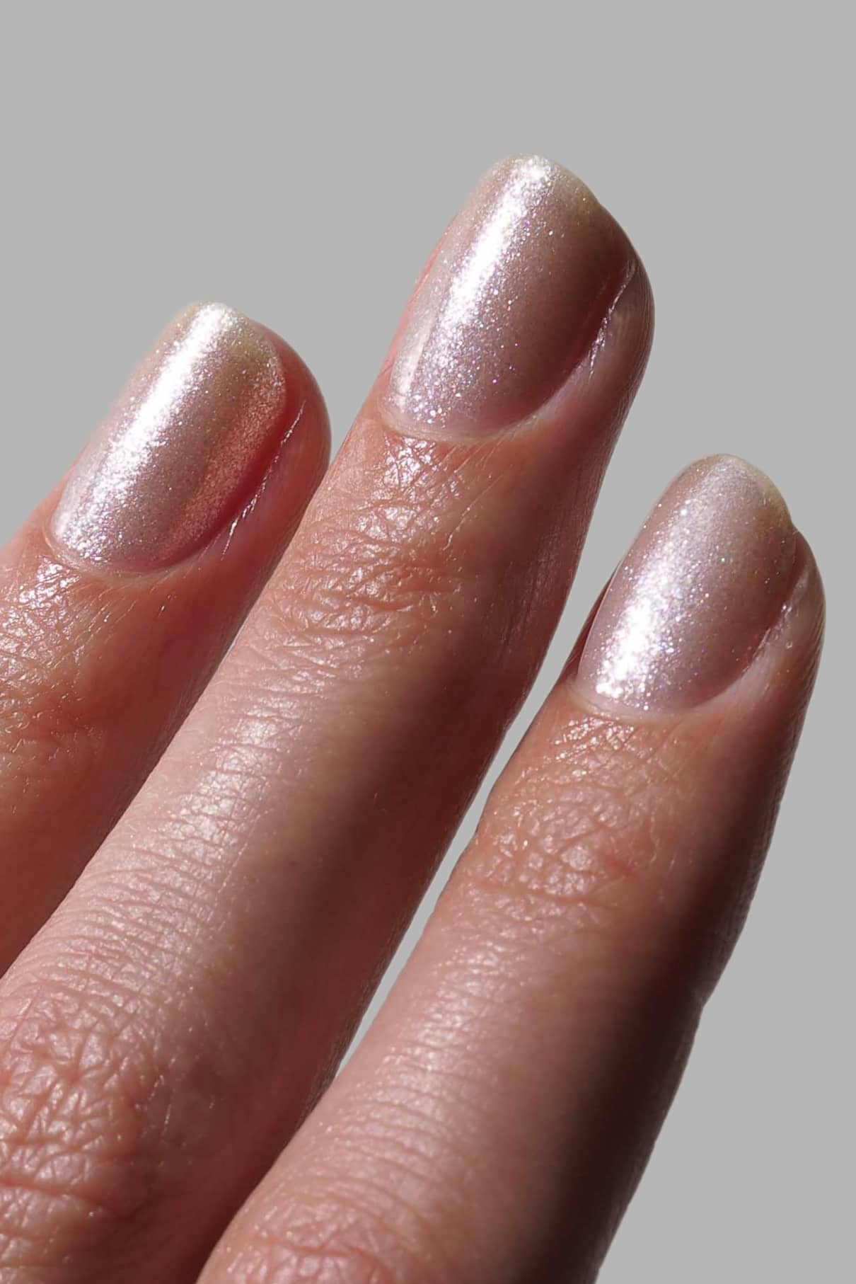 Nahaufnahme von drei Nägeln mit kurzen, sauber gefeilten Spitzen, die mit Nail Highlighter, einer hellen, schimmernden Nagelfarbe, vor einem schlichten grauen Hintergrund lackiert sind.