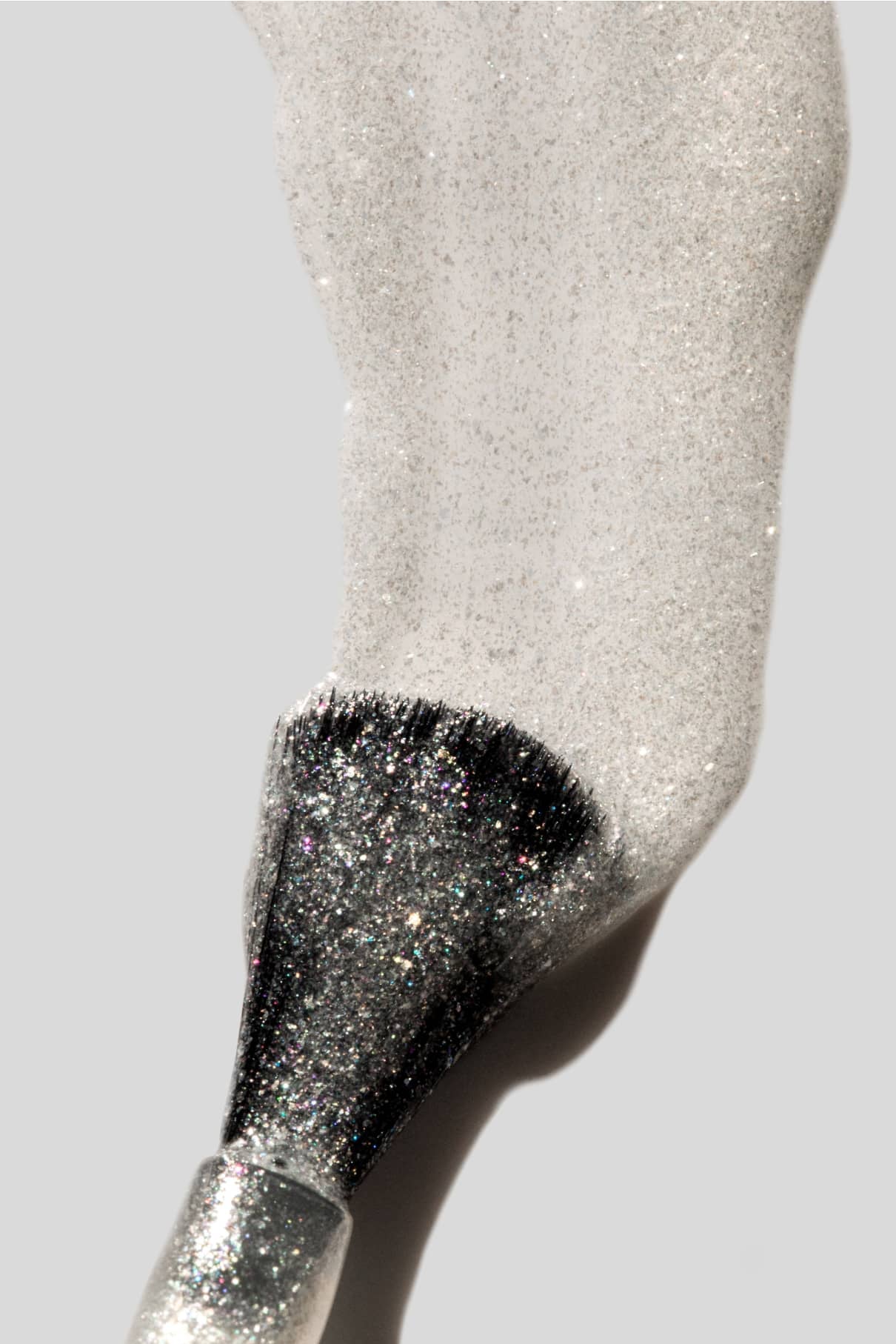 Ein mit feinem, silbernem Glitterpulver bestäubter Nail Highlighter-Pinsel ruht auf einem hellgrauen Hintergrund und erinnert an das schimmernde Finish eleganter Nägel.