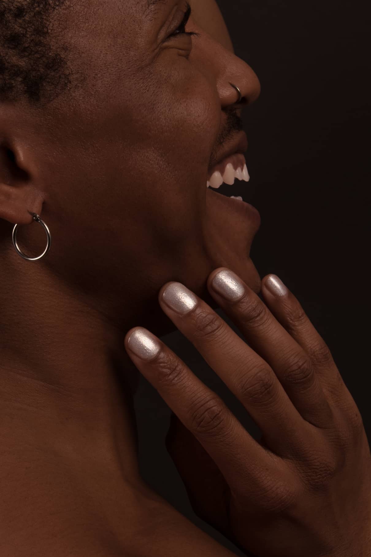 Eine Person mit kurzen Haaren lächelt im Profil und zeigt einen Nasenring und einen Creolenohrring. Ihre Hand, deren Nägel mit dem schimmernden Finish von Nail Highlighter glänzen, berührt ihr Gesicht.