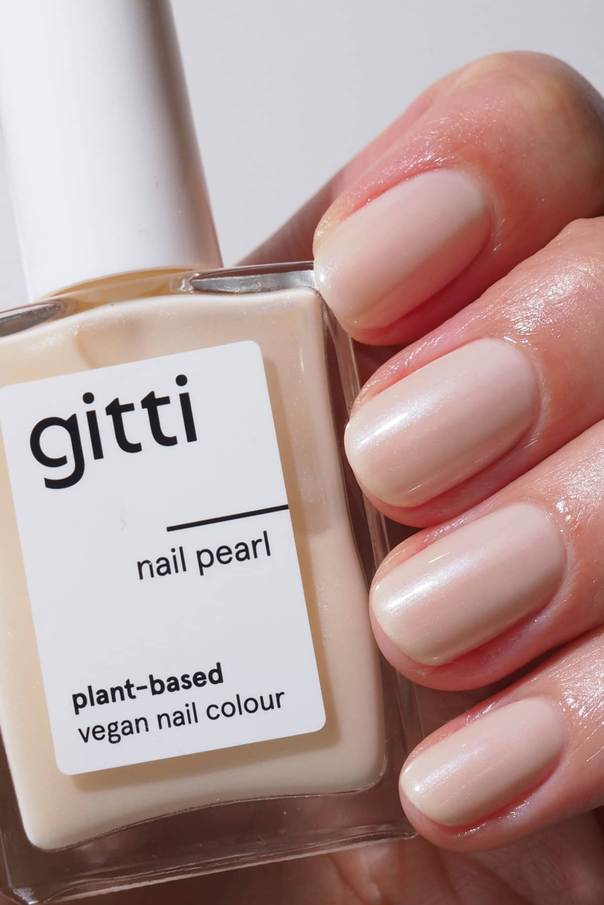 Eine Hand mit manikürten Nägeln hält Nail Pearl, einen pflanzlichen, veganen Nagellack in einem hellen Nude-Ton mit schimmernden Glitzerpartikeln für einen subtilen, halbtransparenten Glanz.