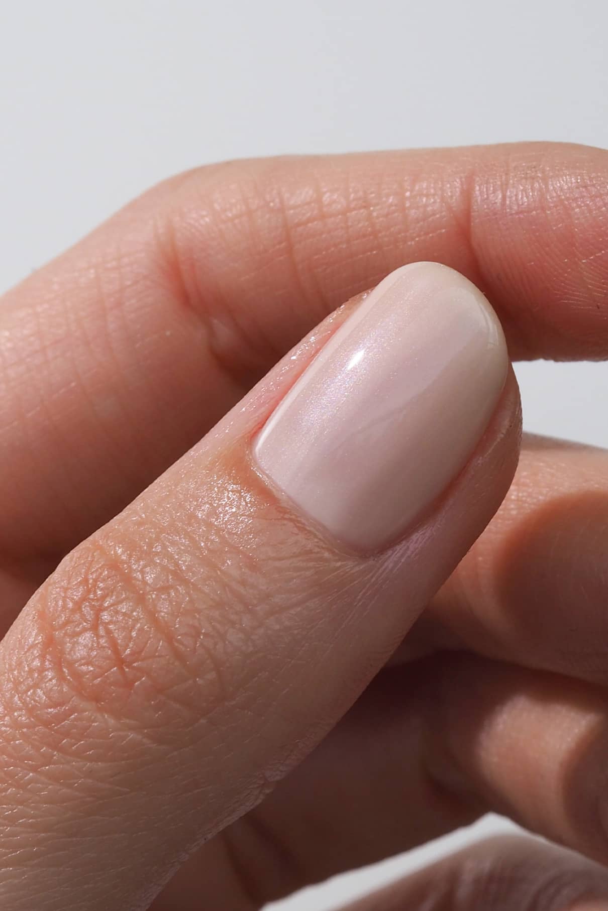 Die Nahaufnahme zeigt den Daumen einer Person mit einem sauber geschnittenen Nagel, der mit Nail Pearl lackiert ist, einer halbtransparenten, hellen Nude-Creme mit schimmernden Glitzerpartikeln.