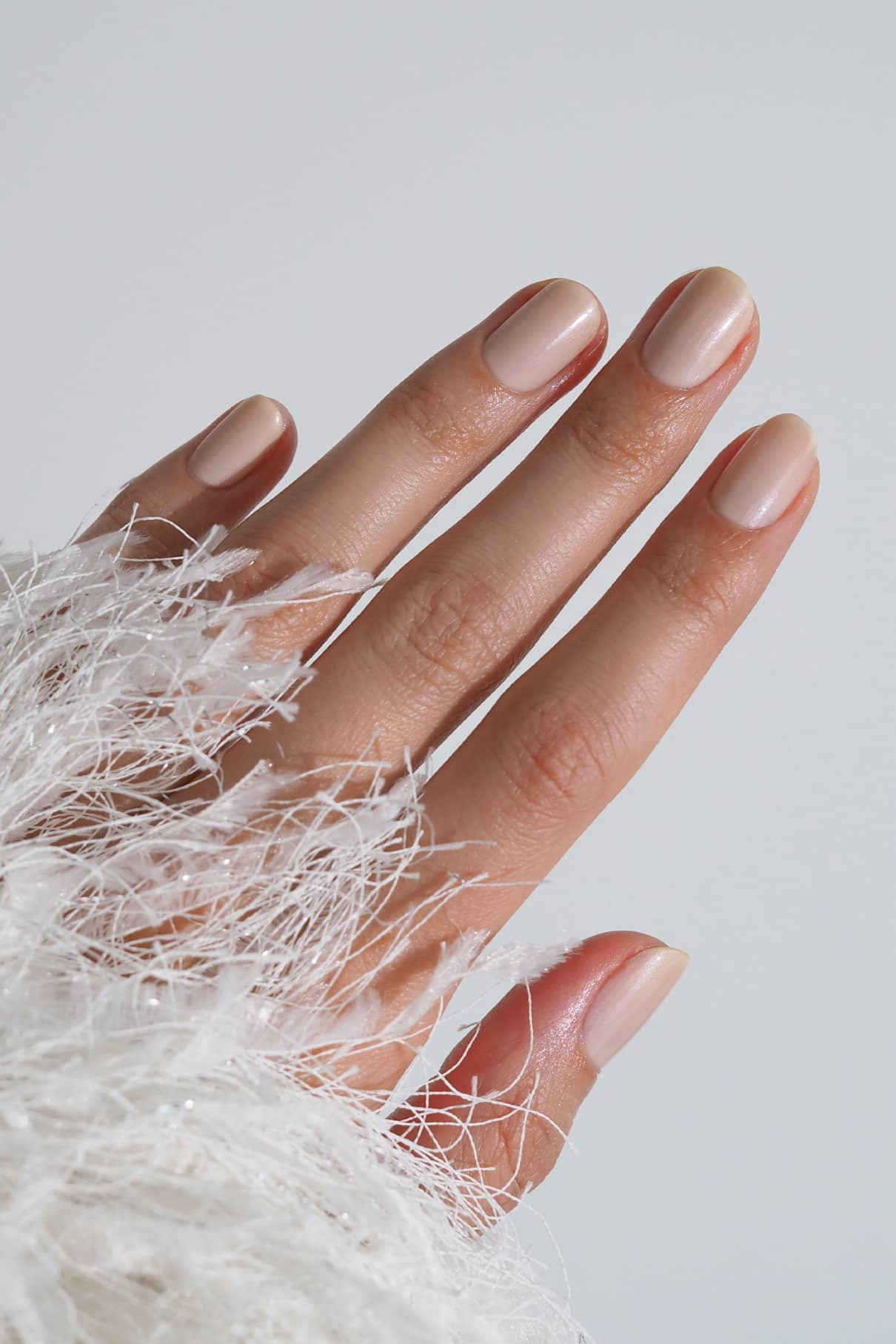 Eine Hand mit kurzen, sauber manikürten, cremefarbenen Nägeln, die mit Nail Pearl Lack lackiert sind, ruht vor einem unifarbenen Hintergrund, der teilweise in strukturierten weißen Stoff gehüllt ist. Die halbtransparente Formel verleiht einen subtilen Glanz mit schimmernden Glitzerpartikeln.