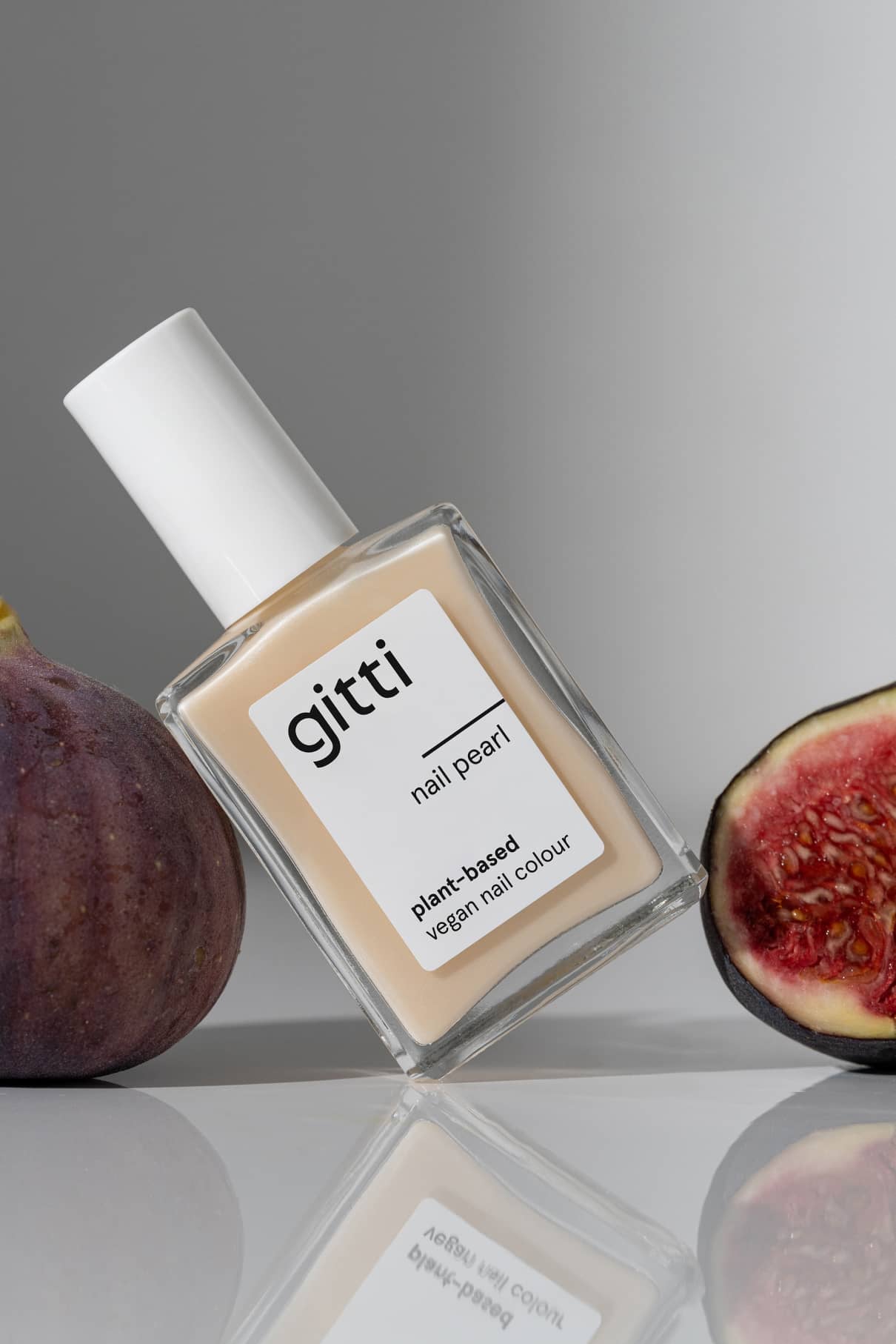 Eine Flasche Nail Pearl pflanzlicher veganer Nagellack mit schimmerndem Glitter steht aufrecht zwischen einer ganzen und einer halbierten Feige auf einer spiegelnden Oberfläche.