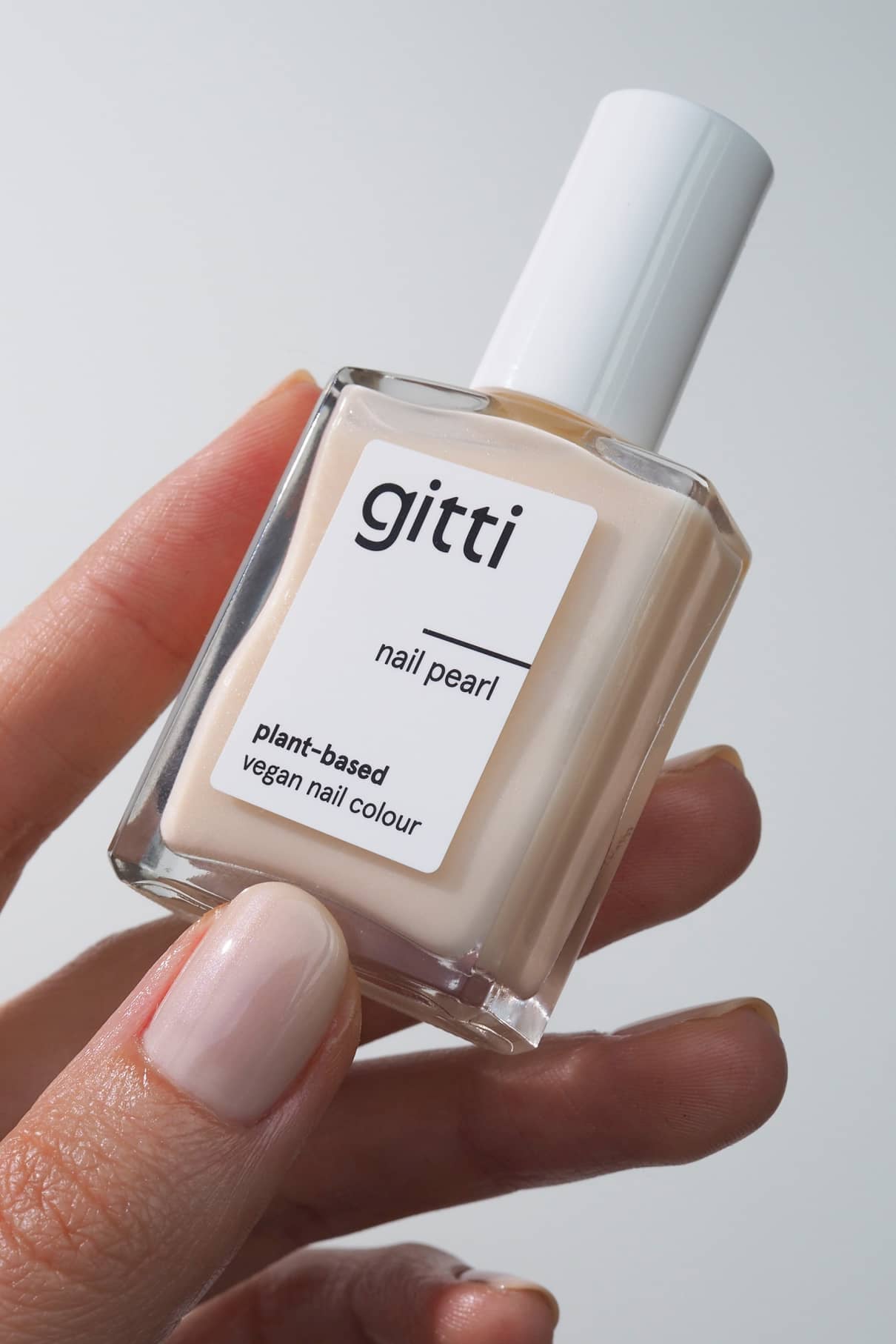 Eine Hand hält eine Flasche Nail Pearl, eine pflanzliche, vegane Nagelfarbe in blassbeiger Creme mit halbtransparenten, schimmernden Glitzerpartikeln für ein dezentes, leuchtendes Finish.