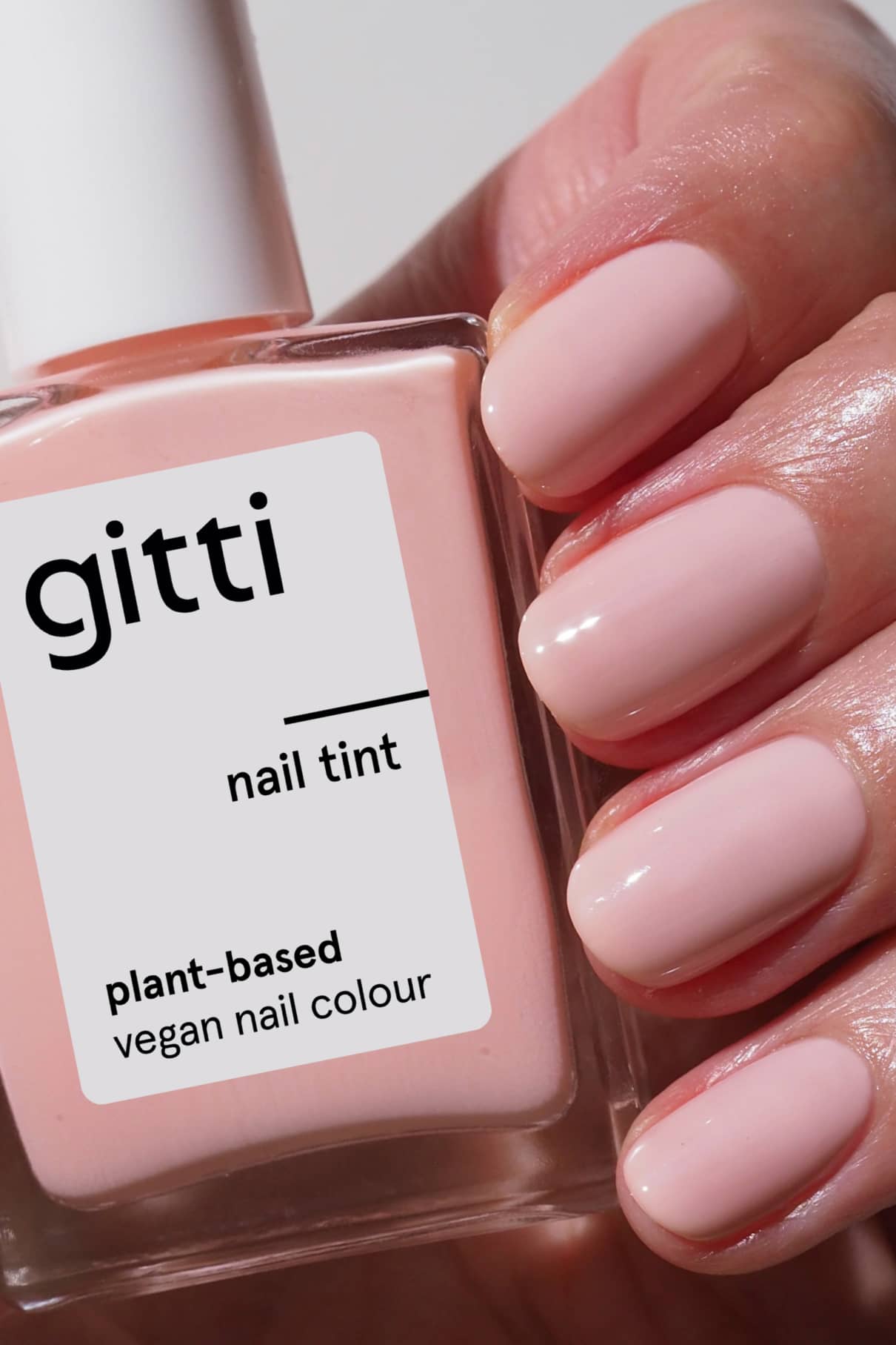 Eine Hand mit sauber manikürten Nägeln in einem milchigen Blassrosa hält eine Flasche Gitti Nail Tint, die als pflanzliche und vegane Nagelfarbe gekennzeichnet ist.