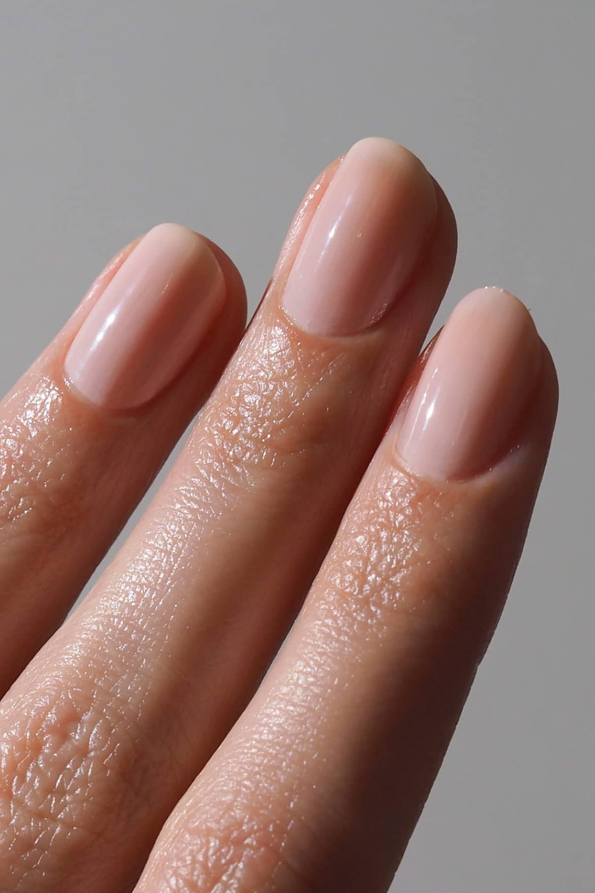 Nahaufnahme von drei Fingern mit sauber manikürten Nägeln, die mit Nail Tint beschichtet sind und einen milchig-zarten Rosaton vor einem unifarbenen Hintergrund aufweisen.