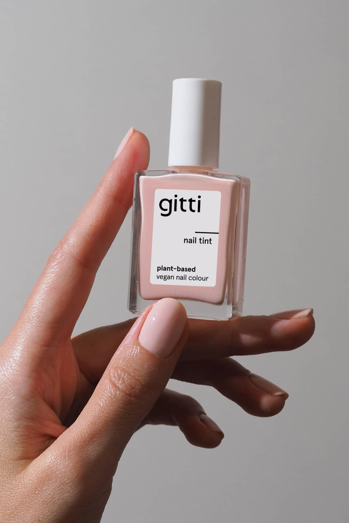 Eine Hand mit nackten Nägeln in einem milchig-zarten Rosaton hält eine Flasche Nail Tint, die als vegane Nagelfarbe auf pflanzlicher Basis gekennzeichnet ist.