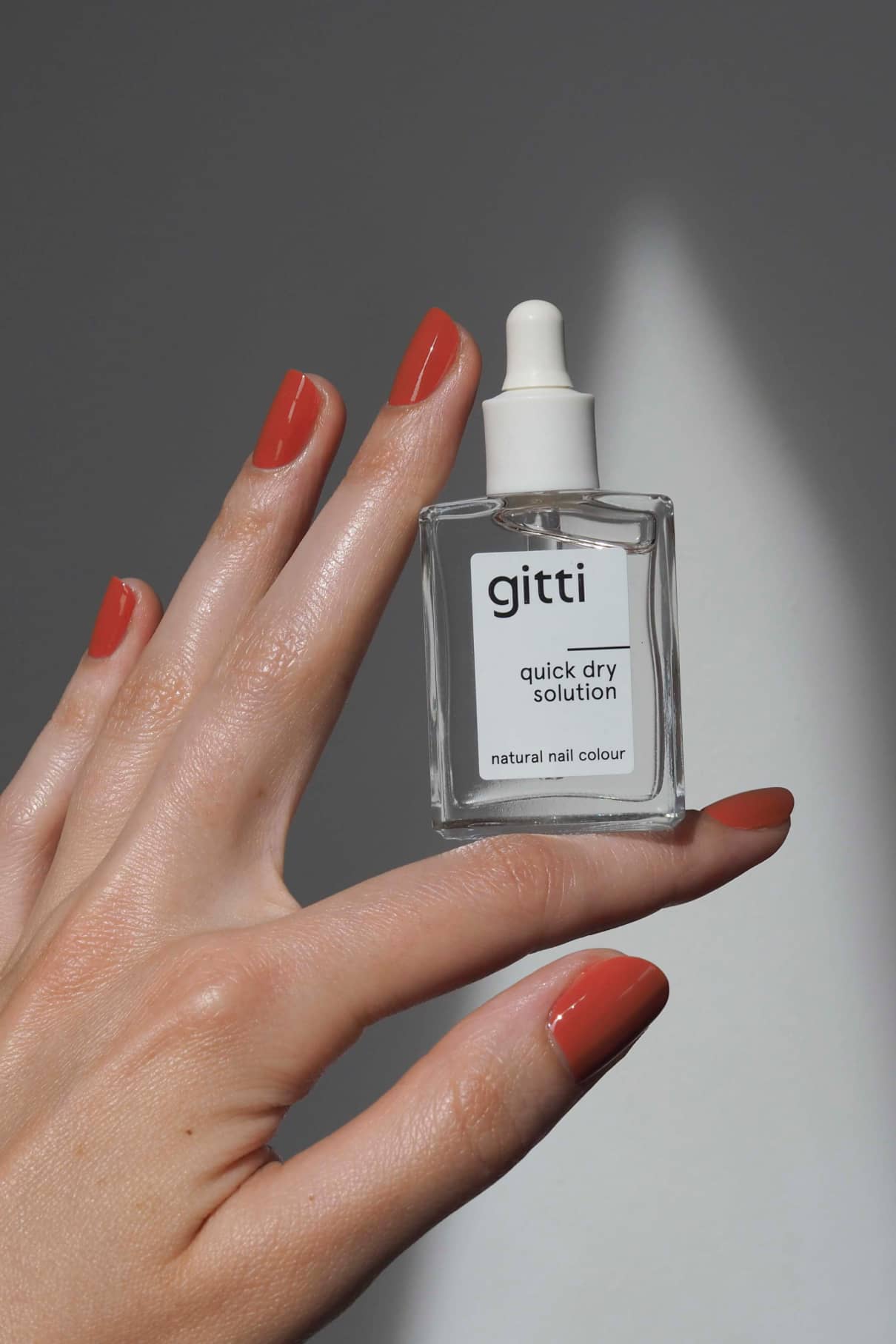 Eine Hand mit lackierten Nägeln hält eine Flasche mit Quick Dry Solution für schnelle Nagellacktrocknung vor einem neutralen Hintergrund.