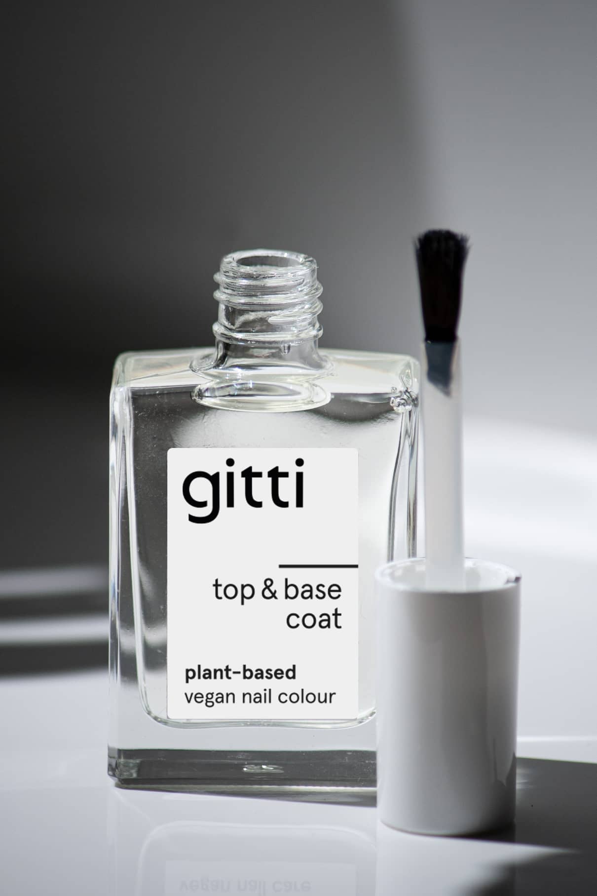Eine offene Klarglasflasche mit der Aufschrift Plant-based Top&Base Coat und einem weißen Pinselapplikator daneben - Ihr 2-in-1-Produkt für zusätzlichen Halt und Glanz.