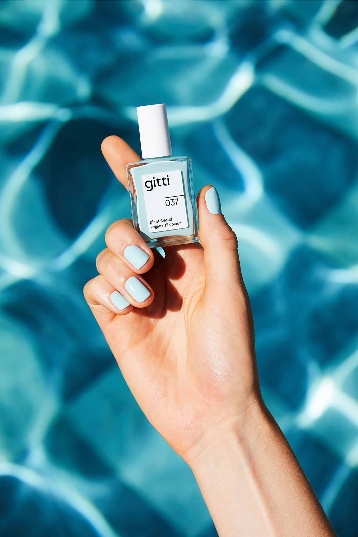 Eine Hand mit hellblauen Nägeln hält Plant Trio + Remover Nagellack vor sonnenbeschienenem blauen Wasser - das perfekte Set für ein makelloses Finish.