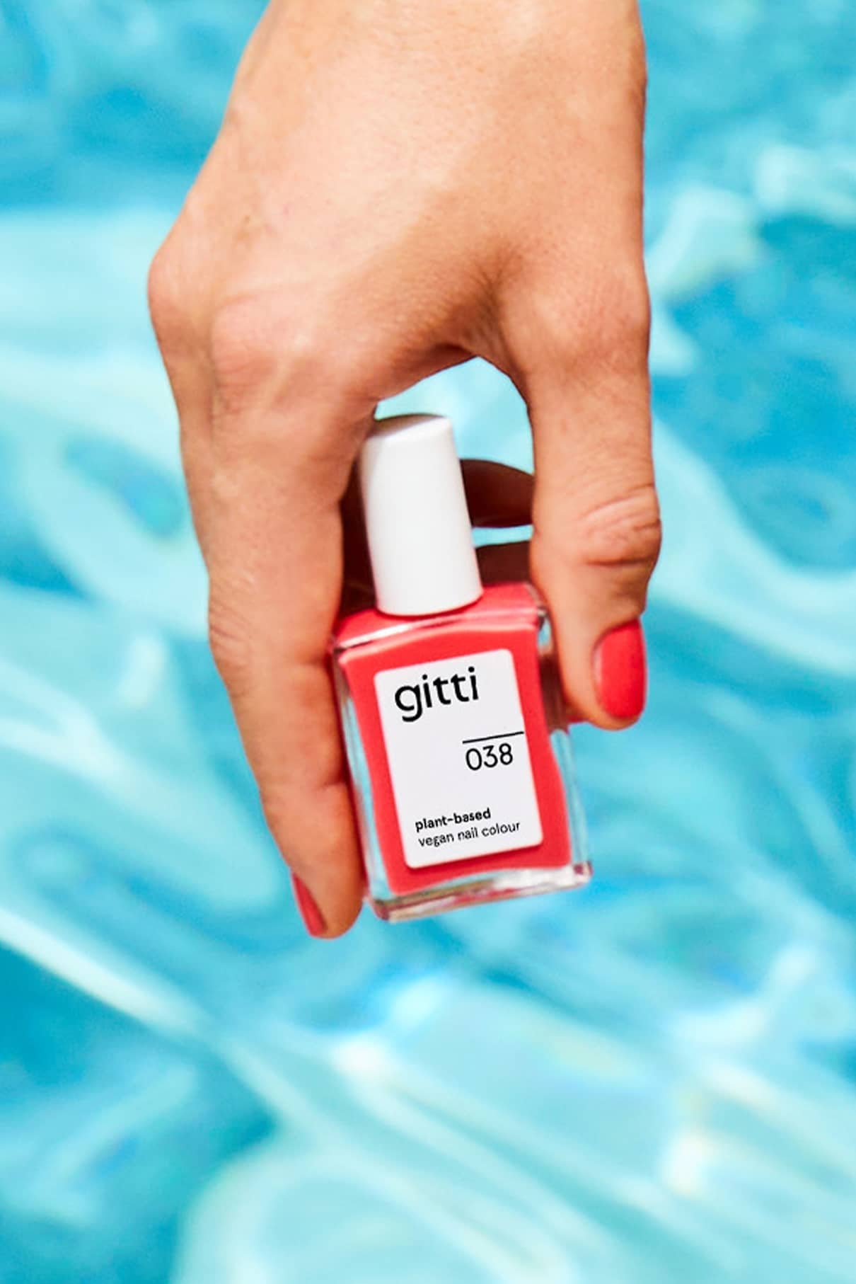 Eine Hand mit lackierten Nägeln hält ein Fläschchen mit gitti no. 038 pflanzliche vegane Nagelfarbe in Korallrot, die vor blauem Hintergrund Wärme und Tiefe ausstrahlt.