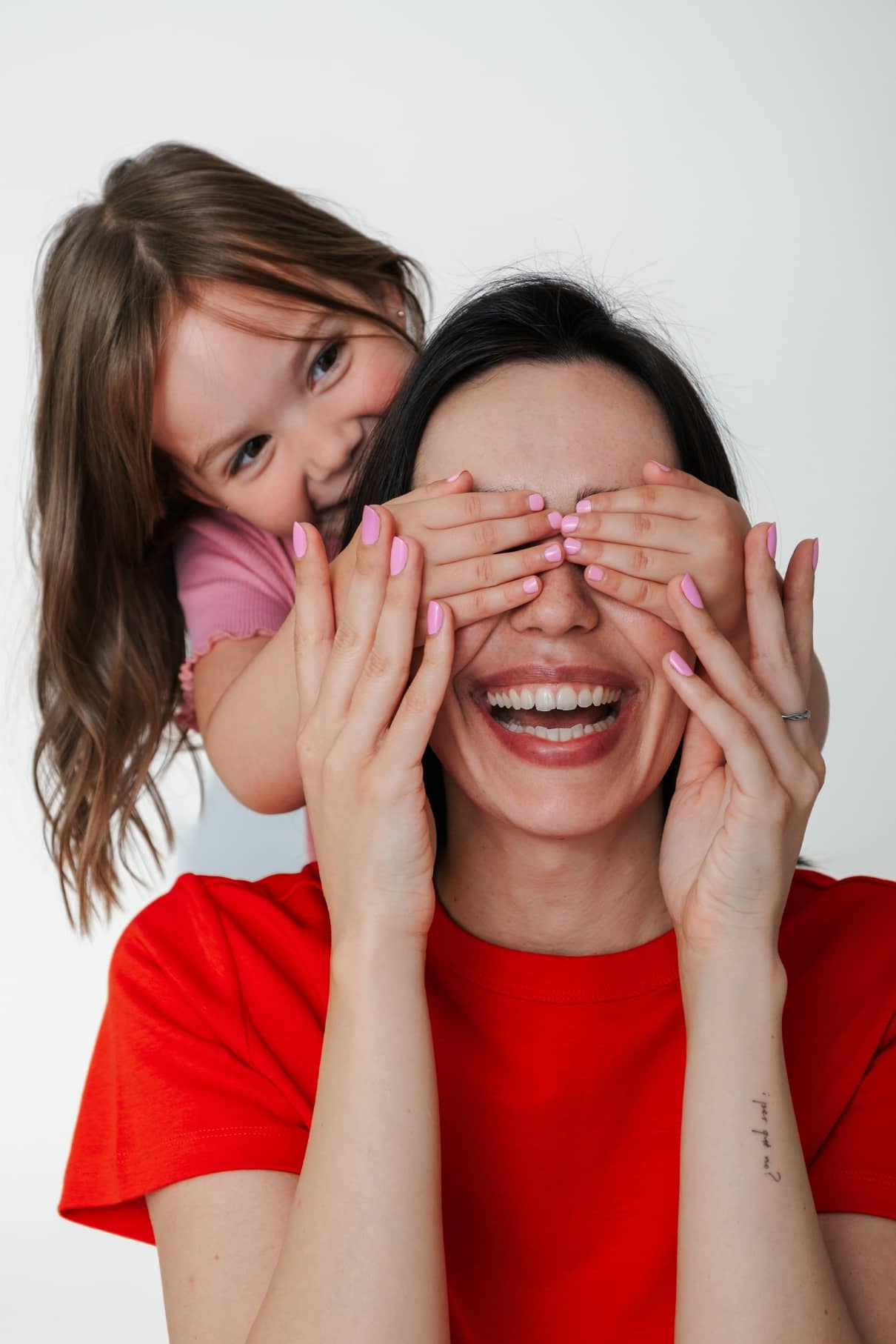Ein junges Mädchen bedeckt spielerisch die Augen einer lächelnden Frau in einem roten Hemd, beide sehen glücklich aus vor einem weißen Hintergrund - perfekt, um das Mini & Me Set von gitti kids hervorzuheben.