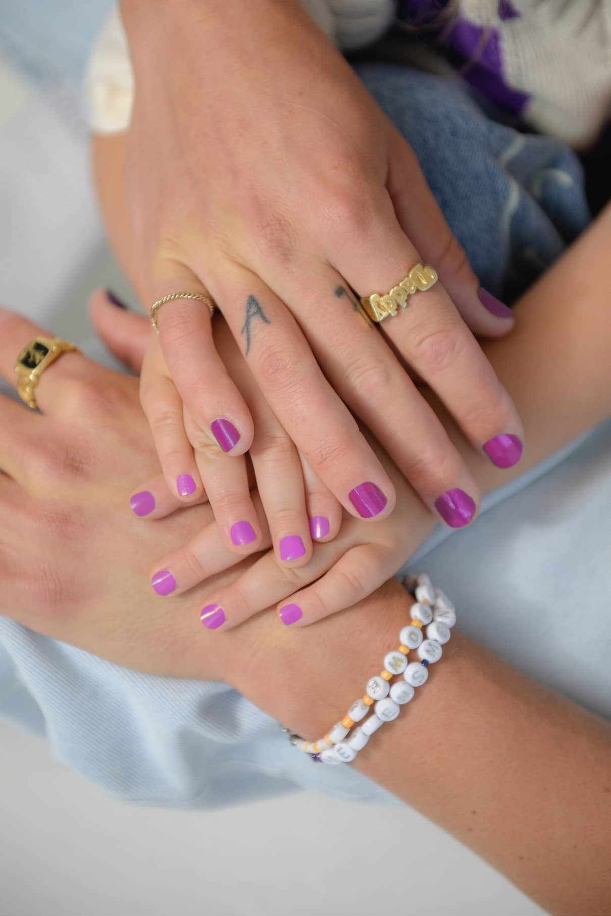 Drei hellhäutige Hände mit lilafarbenem Mini & Me Set-Nagellack sind aneinandergereiht und mit Ringen und Armbändern verziert. Eine Hand ist mit einem kleinen "A" auf dem Finger tätowiert.