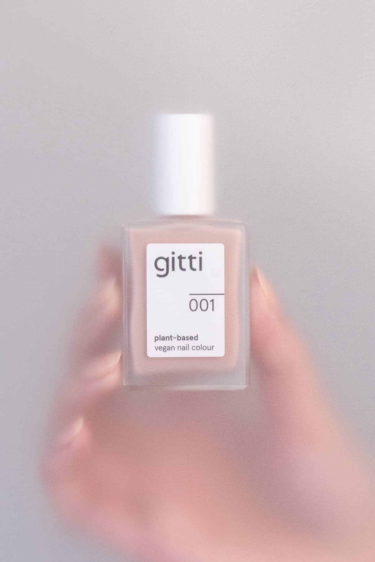 Eine Hand hält eine Flasche mit gitti no. 001 pflanzlicher veganer Nagellack mit weißer Kappe vor einem neutralen Hintergrund, der seinen hellbeigen Farbton hervorhebt.