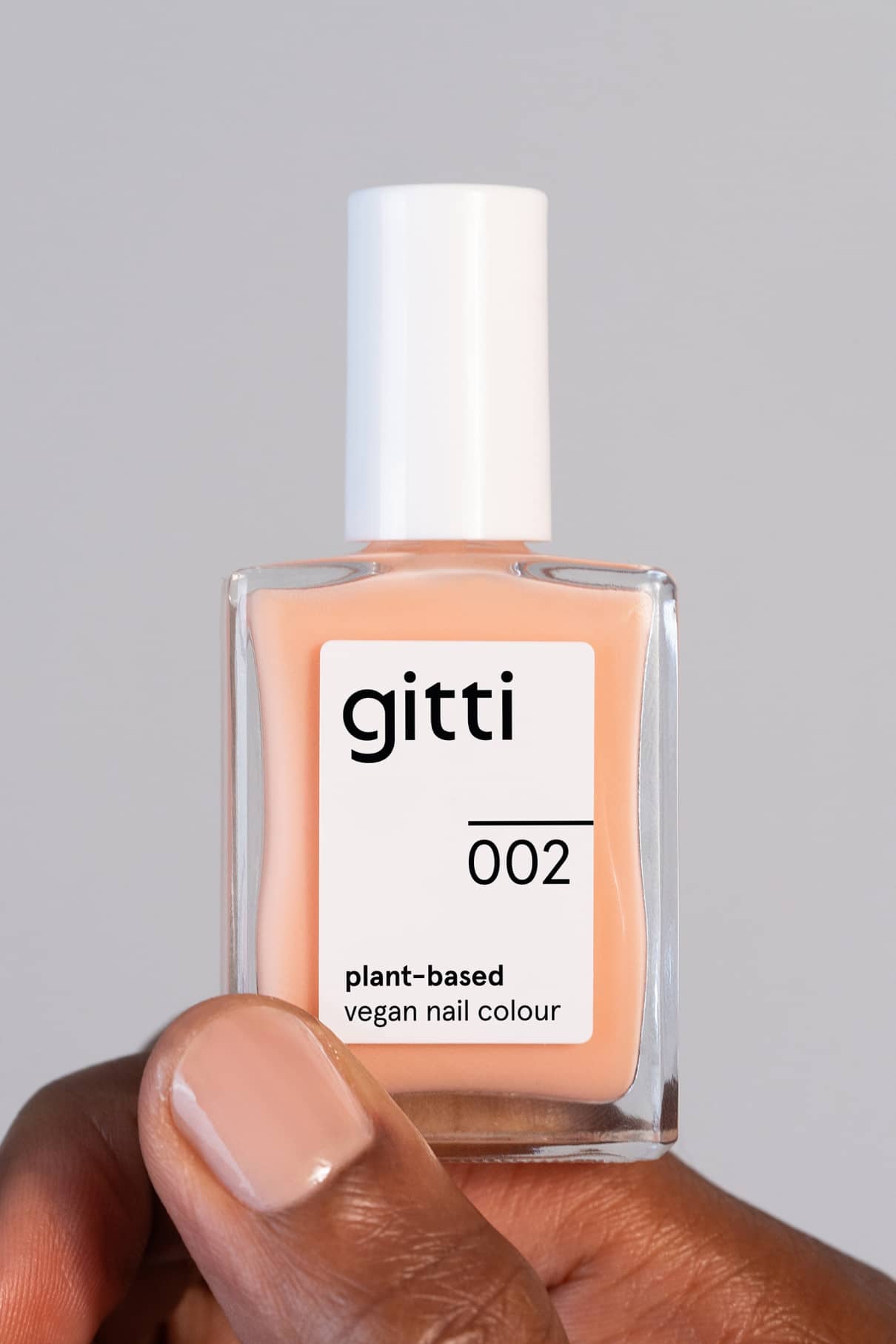 Eine Hand hält eine Flasche mit gitti no. 002 pflanzliche vegane Nagelfarbe. Die Nägel sind in einem Nude-Ton lackiert, ähnlich dem Pfirsichton, Pantone-Farbe des Jahres 2024.