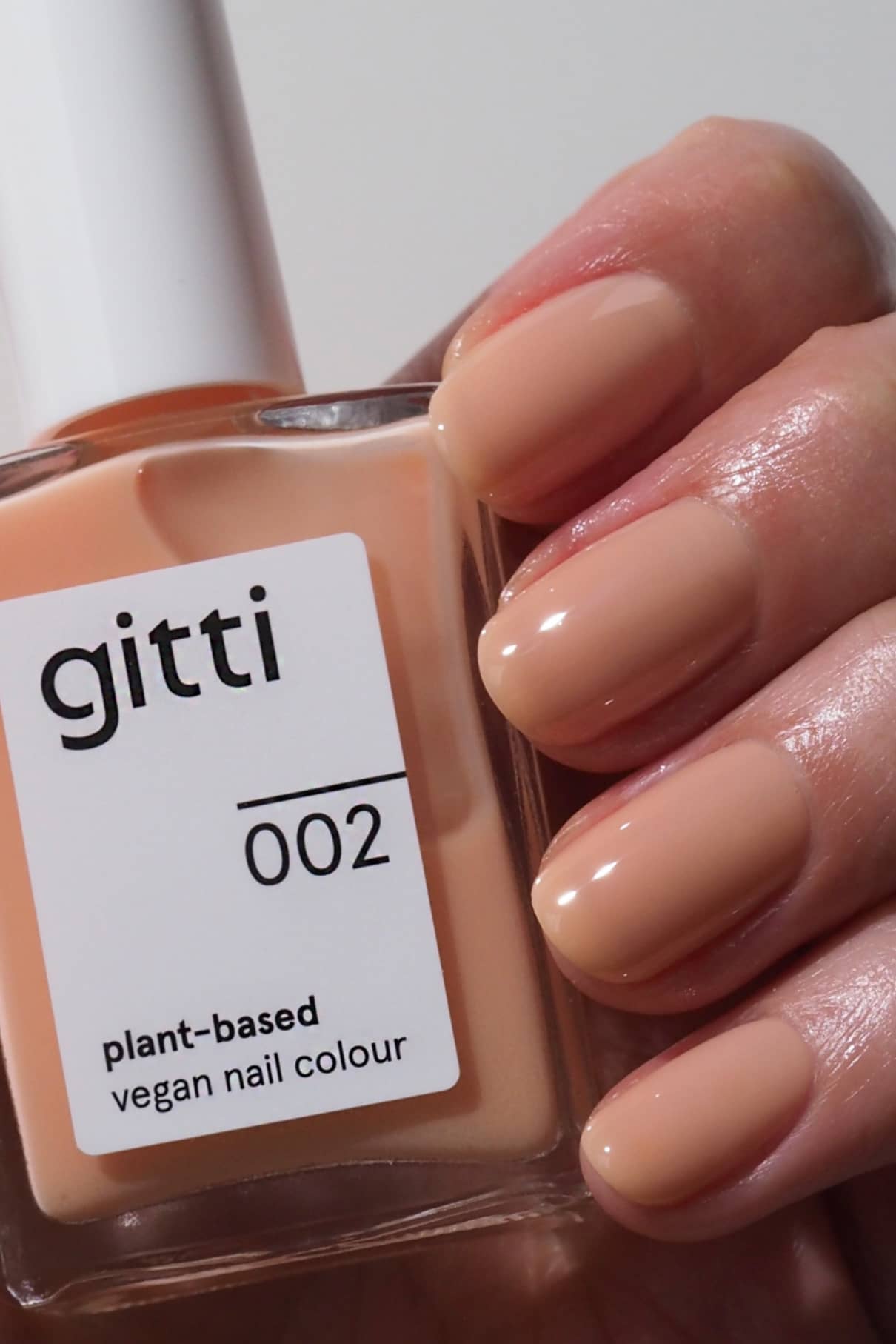 Eine Hand mit kurzen, sauber manikürten Nägeln, die in einem Pfirsichton lackiert sind, der von Pantones Farbe des Jahres 2024 inspiriert ist, hält eine Flasche gitti no. 002, einer veganen Nagelfarbe auf Pflanzenbasis.