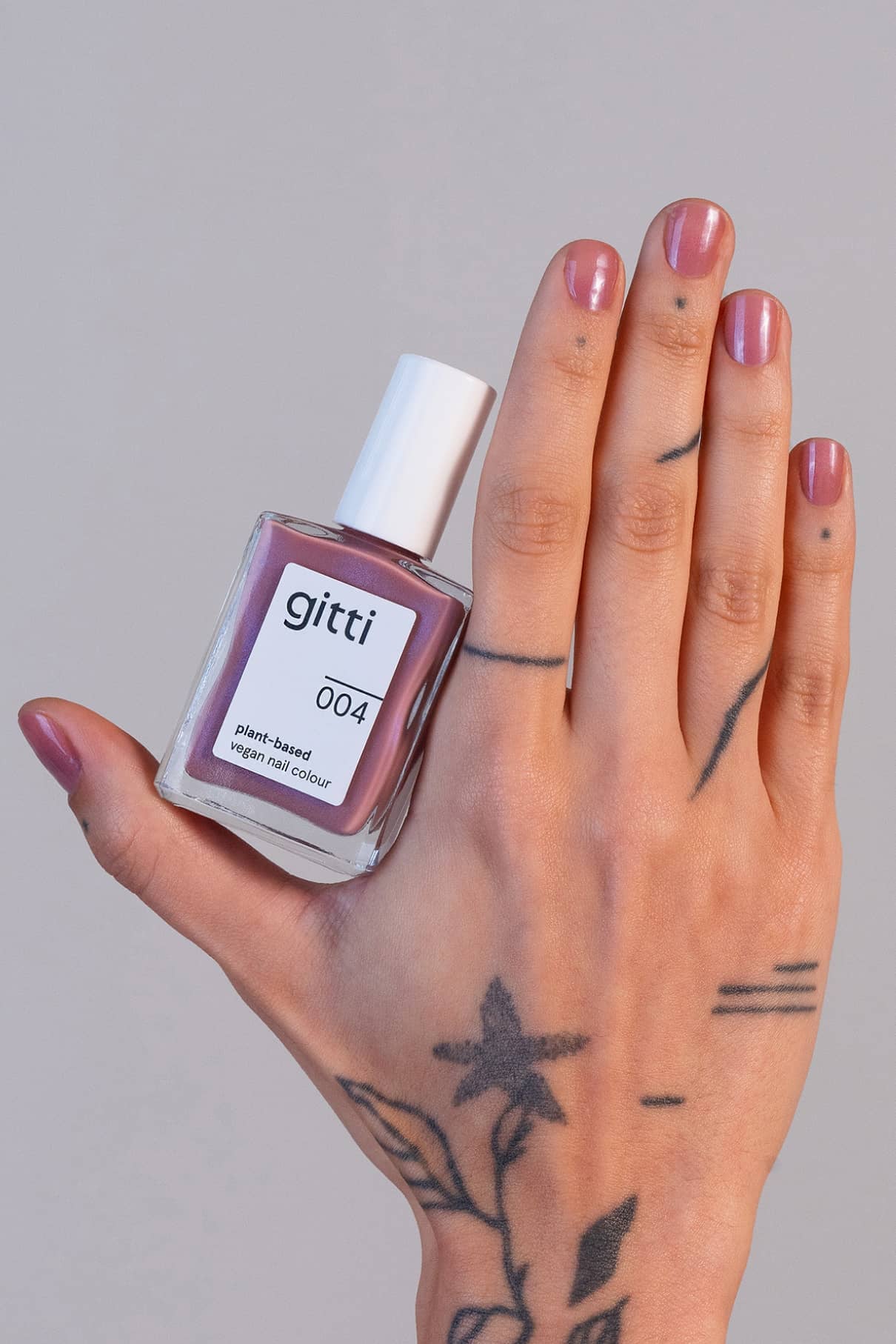Eine tätowierte Hand hält eine Flasche mit gitti no. 004 pflanzliche vegane Nagelfarbe; die Nägel sind in einem passenden seidigen Mauve-Ton lackiert.