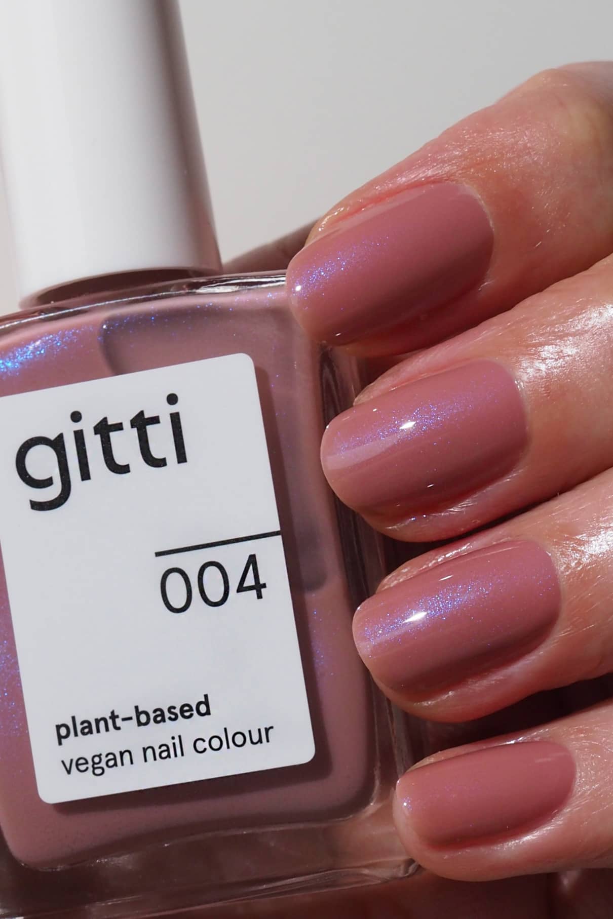 Eine Hand mit kurzen, gepflegten Nägeln, die in seidigem Mauve lackiert sind, hält eine Flasche mit der Aufschrift gitti no. 004 vegane Nagelfarbe auf Pflanzenbasis.