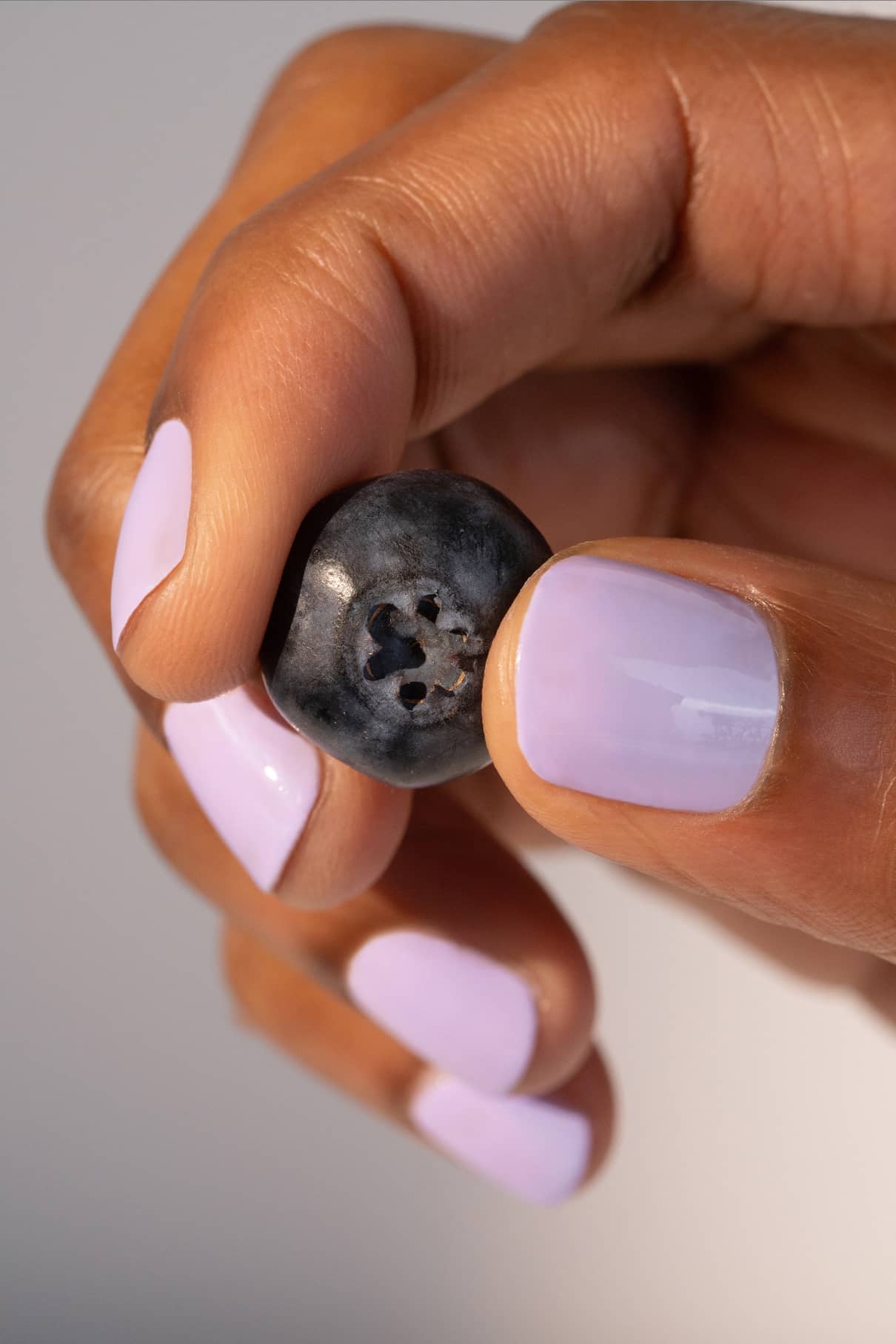 Eine Hand mit gitti no. 005 Nagellack in harmonischem Lila hält eine einzelne Heidelbeere zwischen Daumen und Zeigefinger vor einem neutralen Hintergrund.