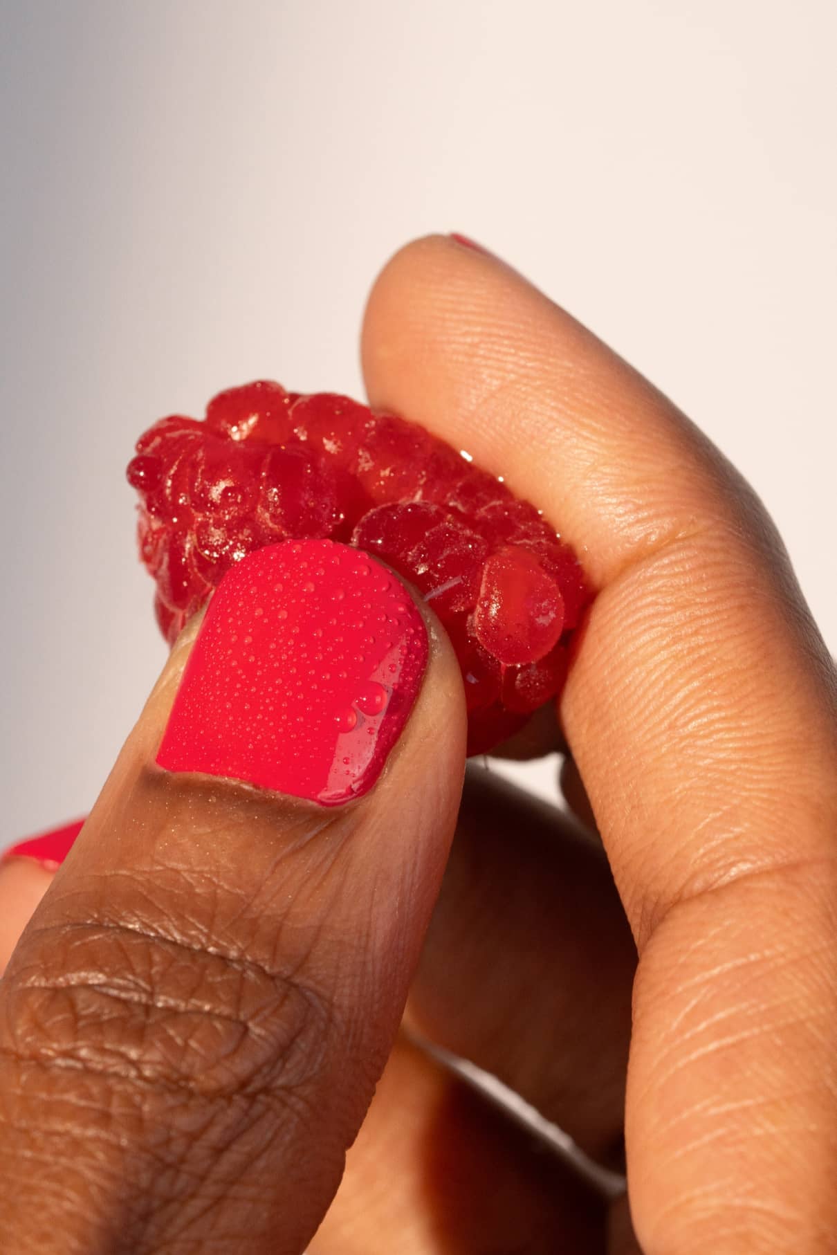 Eine Hand mit glänzendem gitti no. 006 Nagellack, einem leuchtenden Rot mit rosa Unterton, hält eine frische Himbeere zwischen Daumen und Zeigefinger vor einem schlichten Hintergrund.