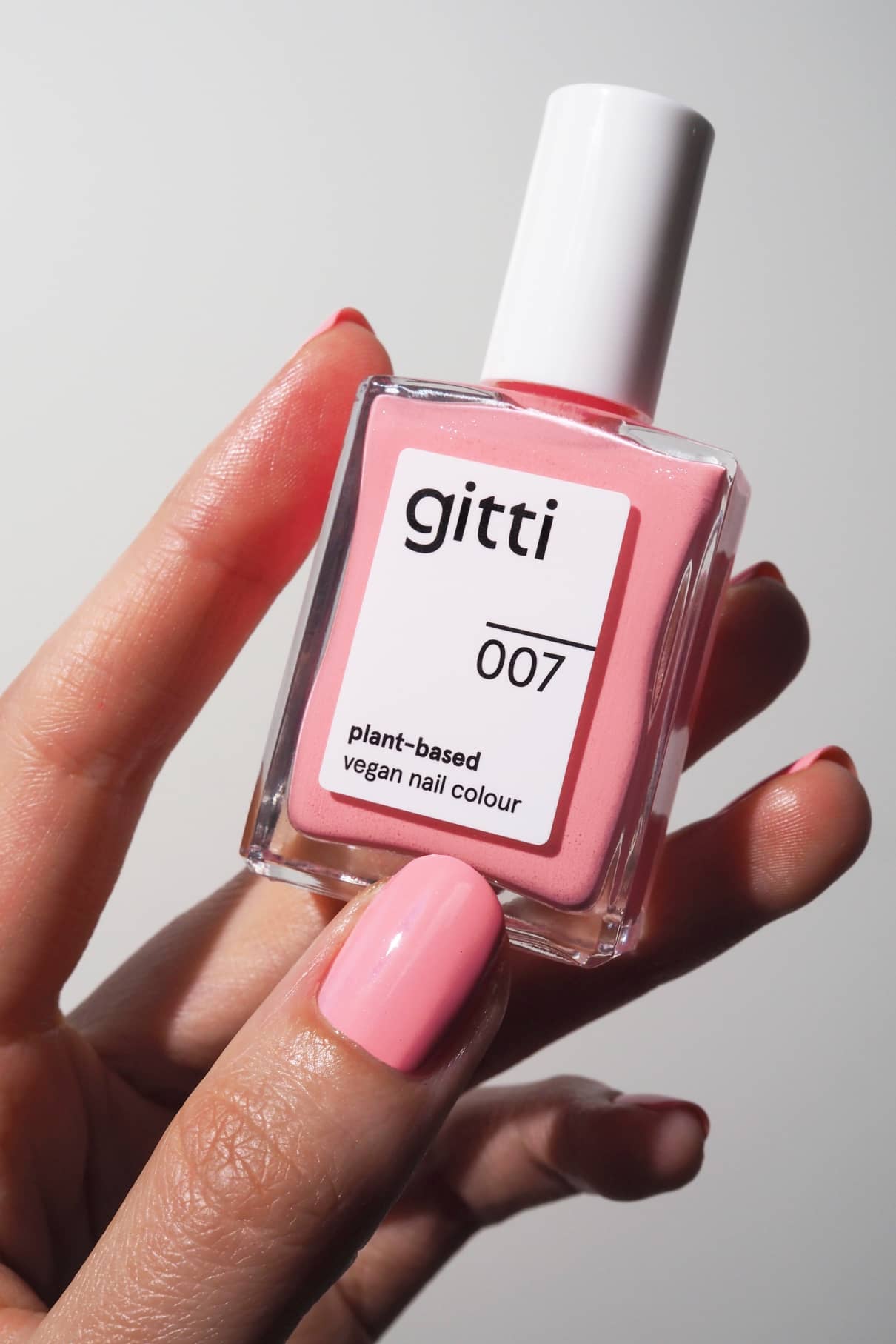 Eine Hand mit rosa lackierten Nägeln hält eine Flasche gitti no. 007 pflanzlichen, veganen Nagellack in einem schimmernden Rosaton, der perfekt zu den Nägeln passt.