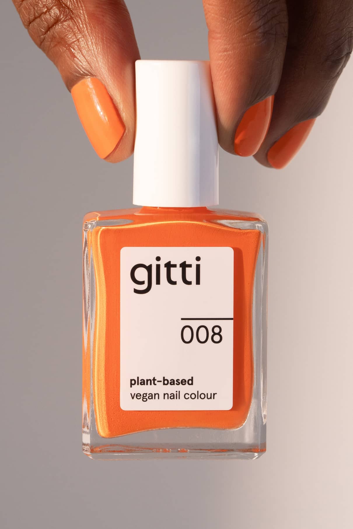Eine Hand mit orange gefärbten Nägeln hält eine Flasche gitti no. 008, einen veganen Nagellack auf Pflanzenbasis, in einem quadratischen Glasbehälter mit weißer Kappe.