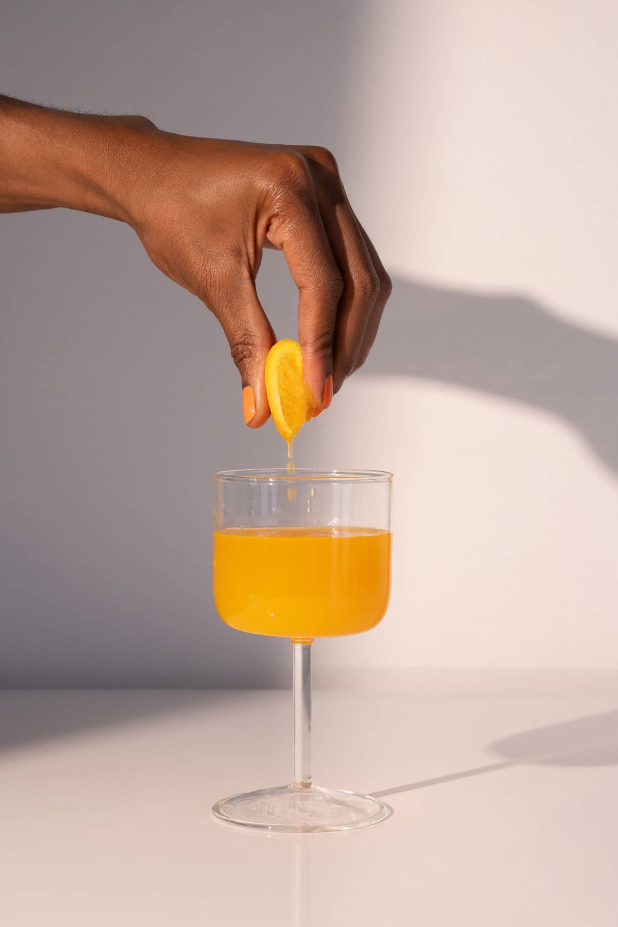 Eine Hand drückt eine Zitronenspalte über einem Glas aus, das mit gitti no. 008, einer orange-gelben Flüssigkeit, auf einer weißen Fläche.