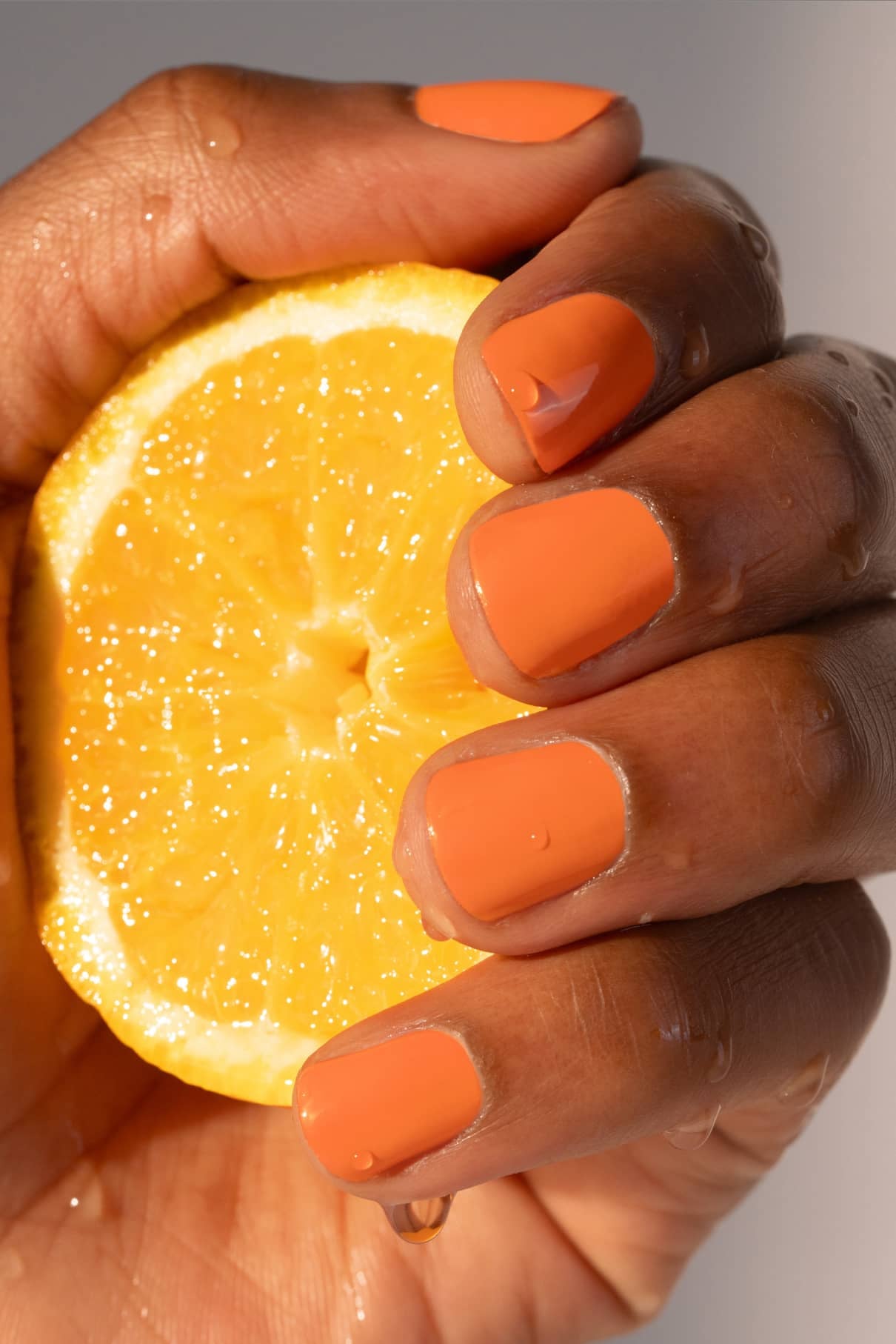 Eine Hand mit gitti no. 008 orange lackierten Nägeln hält eine halbierte Zitrone, auf deren Schale und Frucht Wassertropfen glitzern.