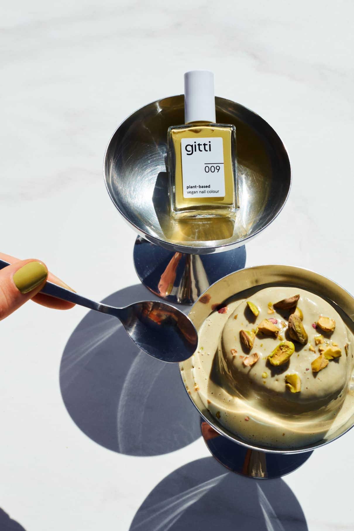 Eine Hand hält einen Löffel neben einer Schale mit Pistaziendessert mit Nüssen; in einer anderen Schale steht eine Flasche gitti no. 009, eine pflanzliche Nagelfarbe in Pistaziengrün mit sanften gelben Untertönen.