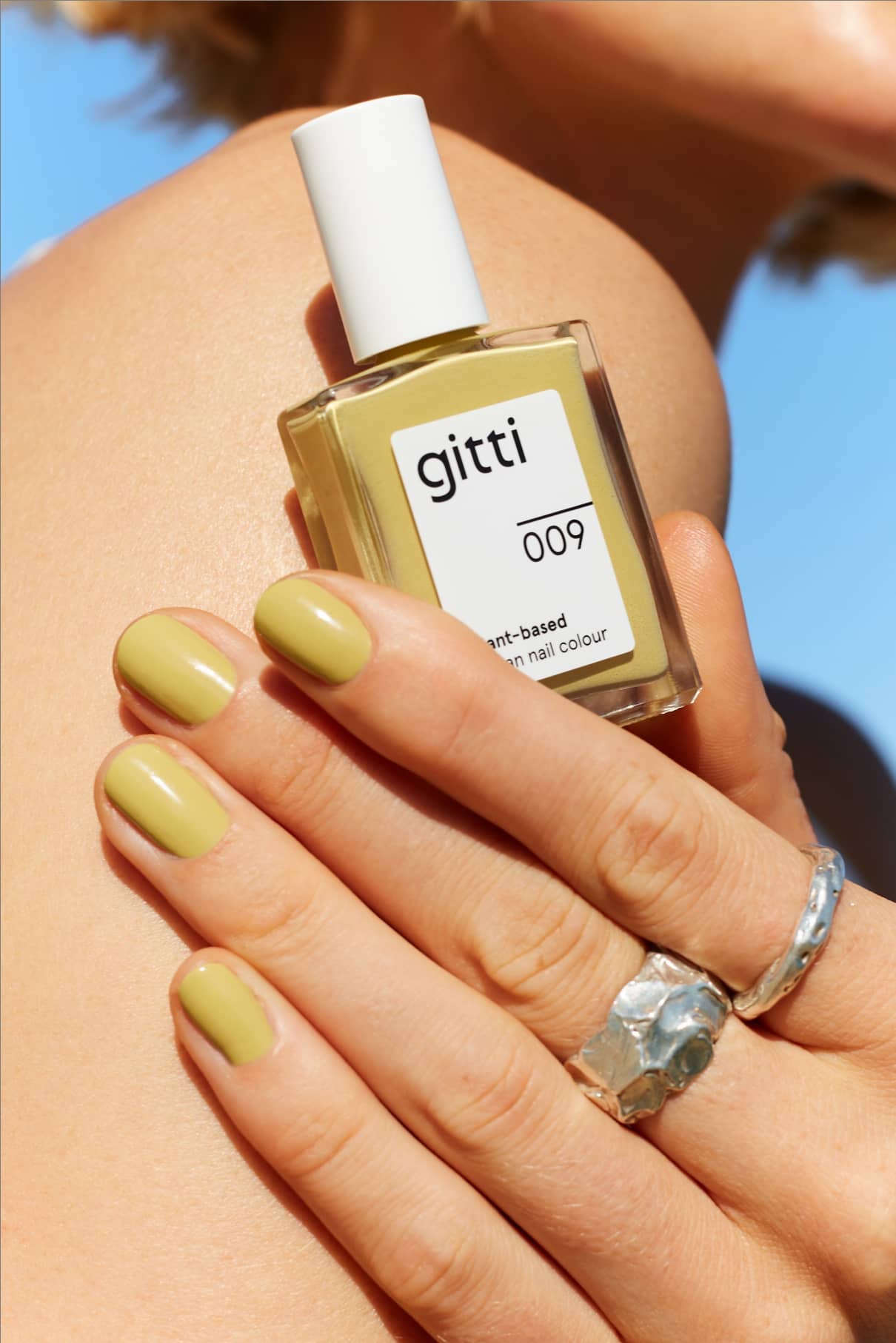 Eine Hand mit pistaziengrünen Nägeln mit zartgelben Untertönen hält eine Flasche gitti no. 009 pflanzlicher Nagelfarbe vor nackter Haut und blauem Himmel.