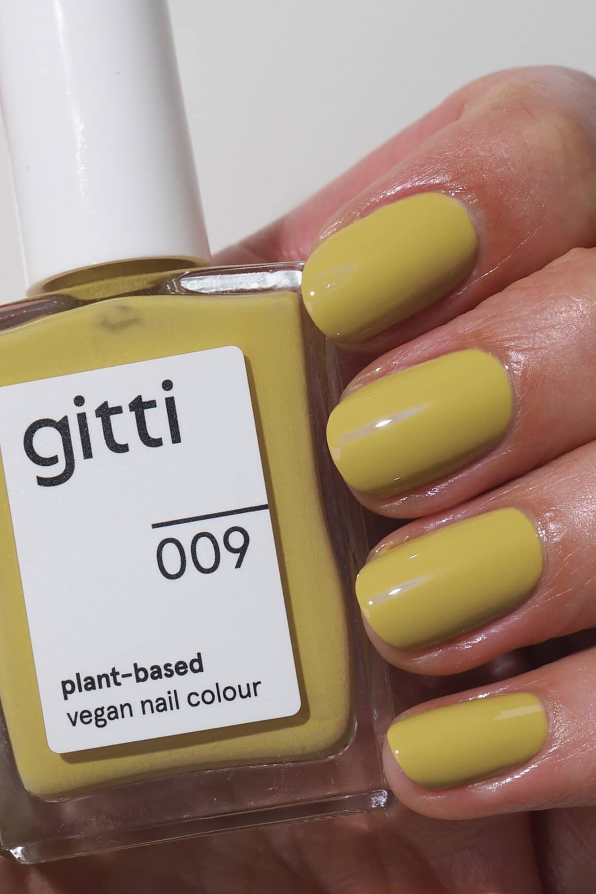 Eine Hand mit kurzen, gepflegten Nägeln, die pistaziengrün mit zartgelben Untertönen lackiert sind, hält eine Flasche gitti no. 009 veganen Nagellack auf Pflanzenbasis.